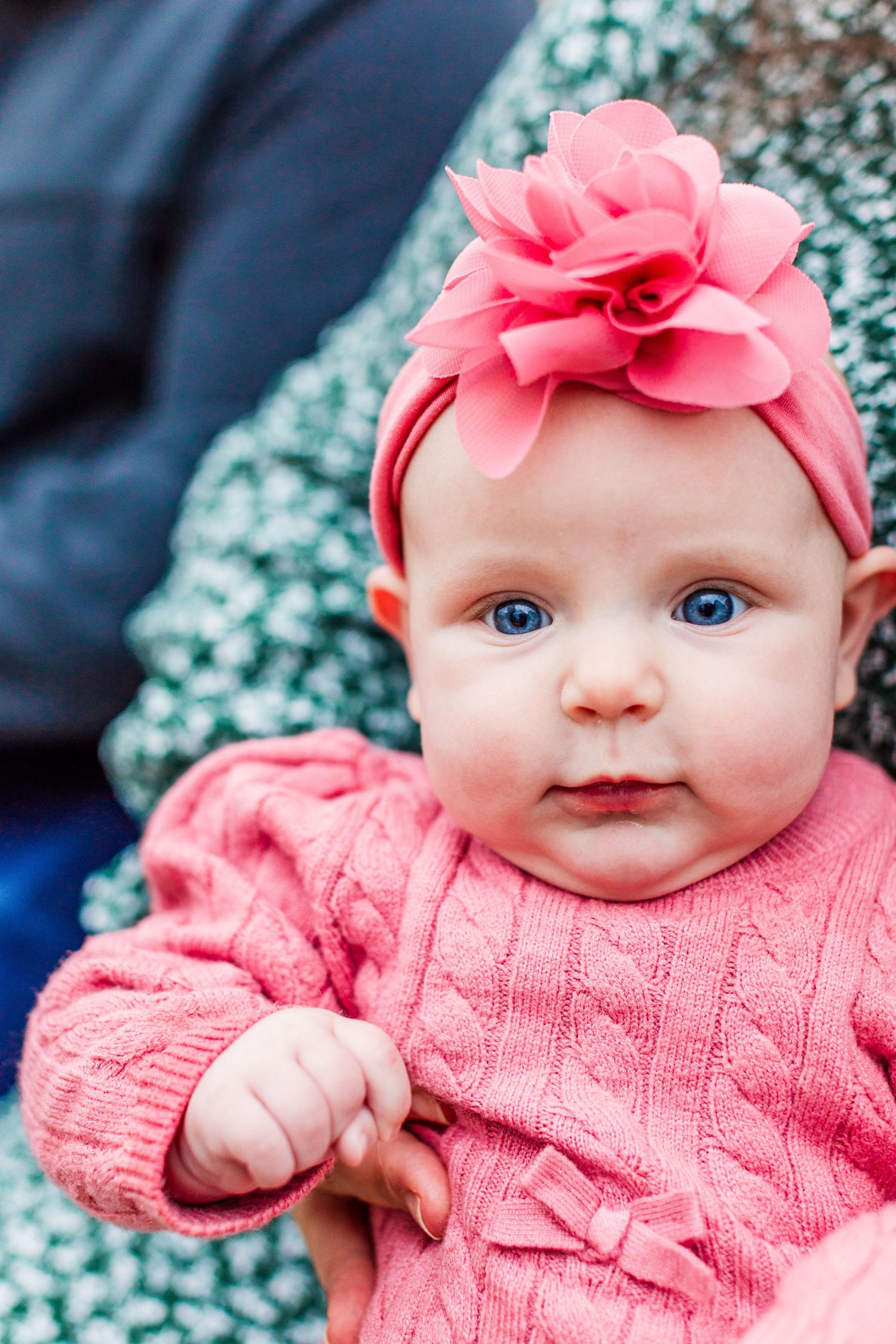 A baby wearing a pink sweater and floral headband smiles at the camera.