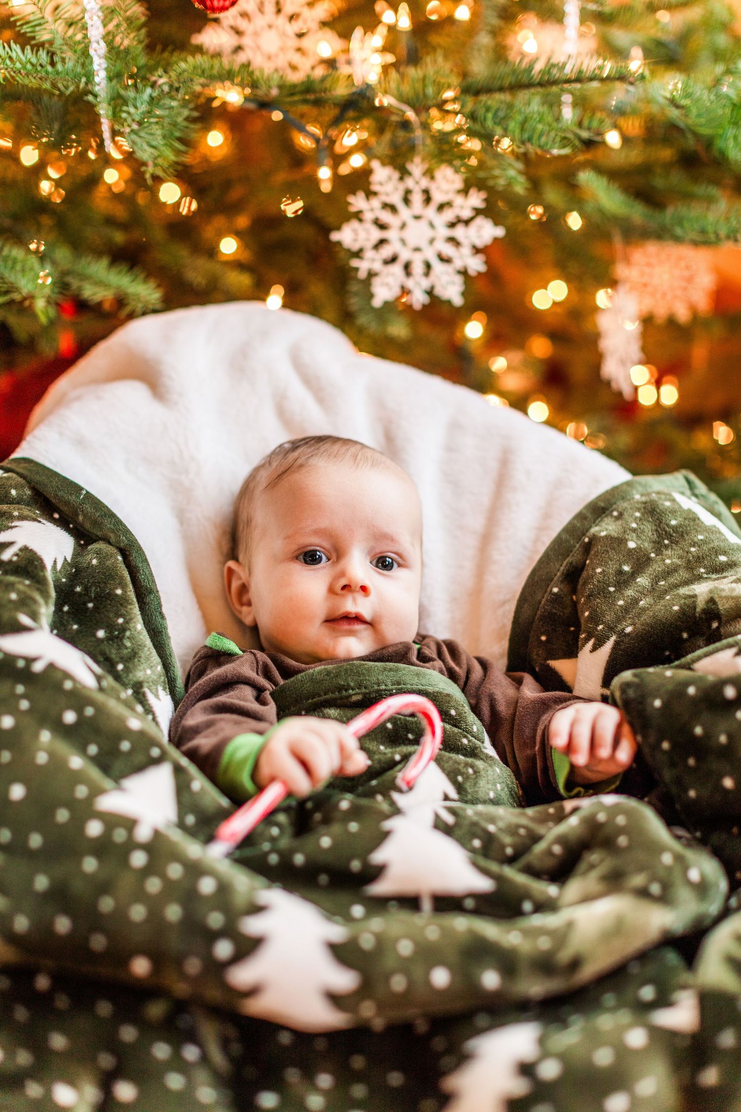 Baby lying under Christmas tree with snowflake decorations and twinkling lights.