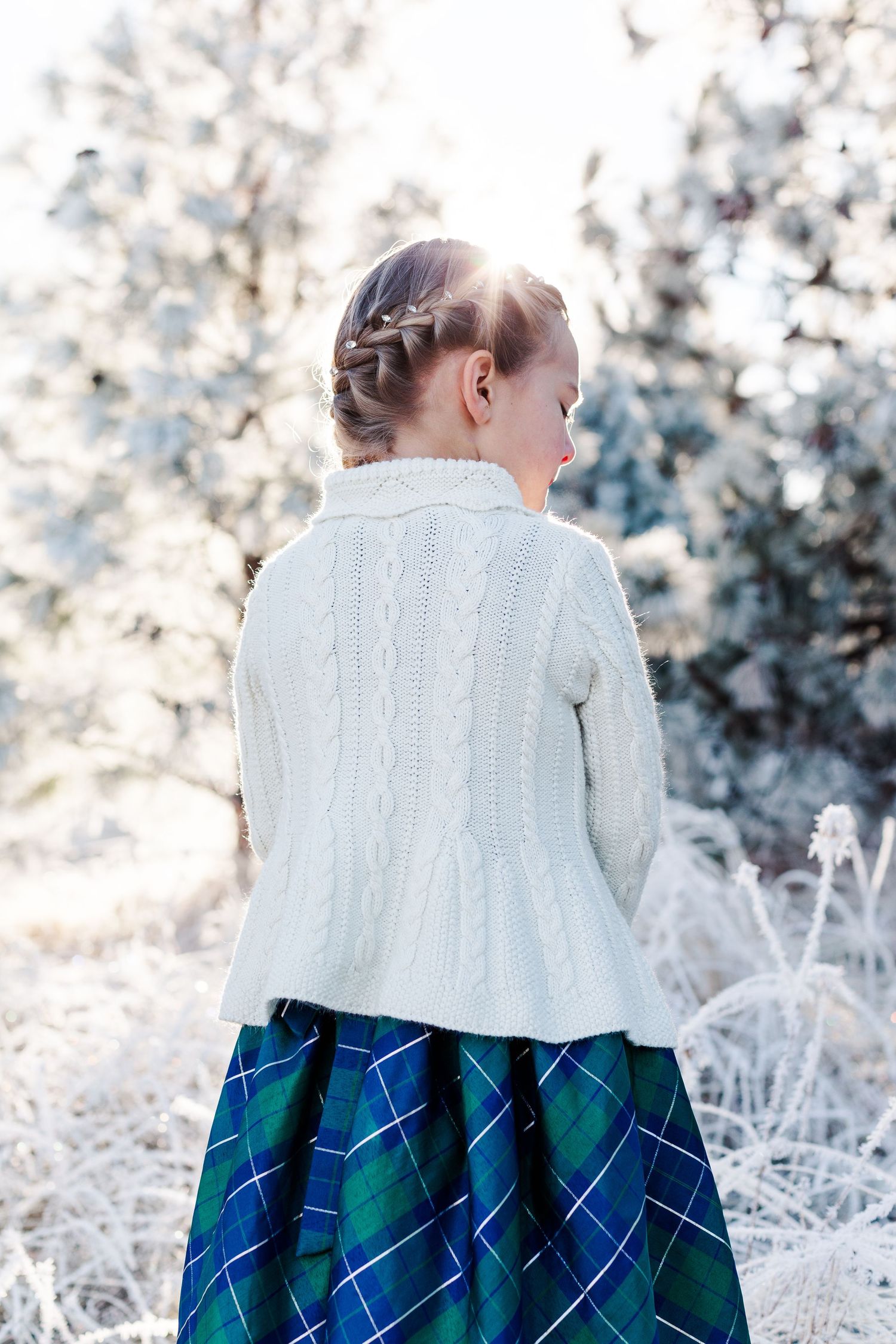 Back view of child in white sweater and plaid skirt against snowy winter backdrop.