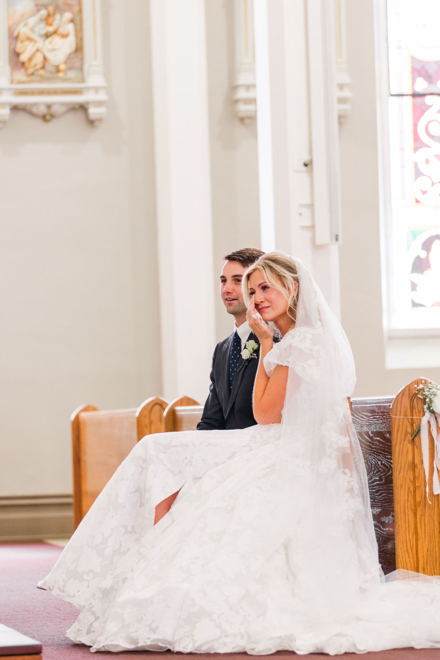 Bride and groom sitting together in a church pew during their wedding ceremony, bride in lace gown with veil.