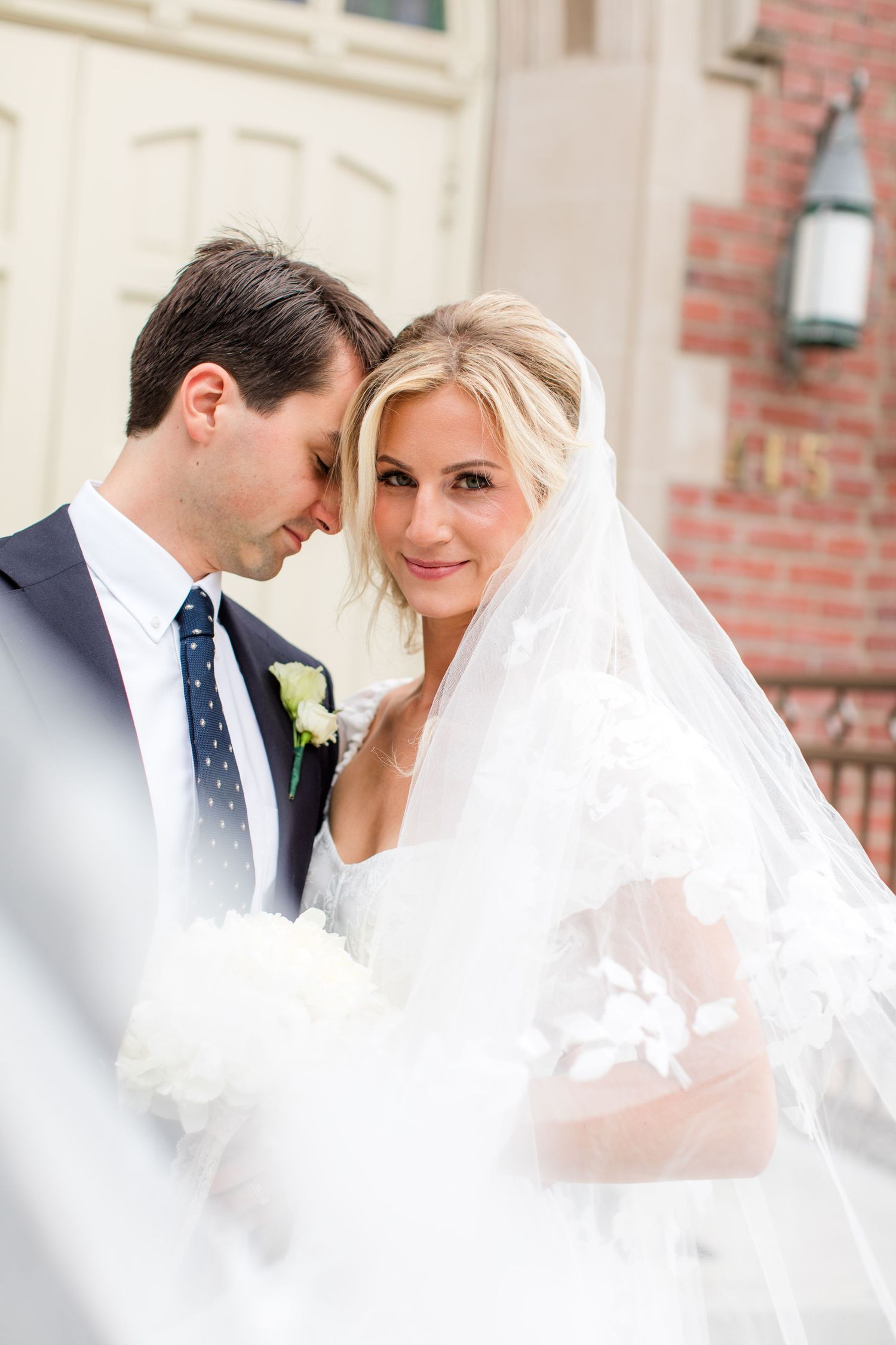 Bride in white veil and groom in navy suit share intimate moment on church steps on their wedding day.