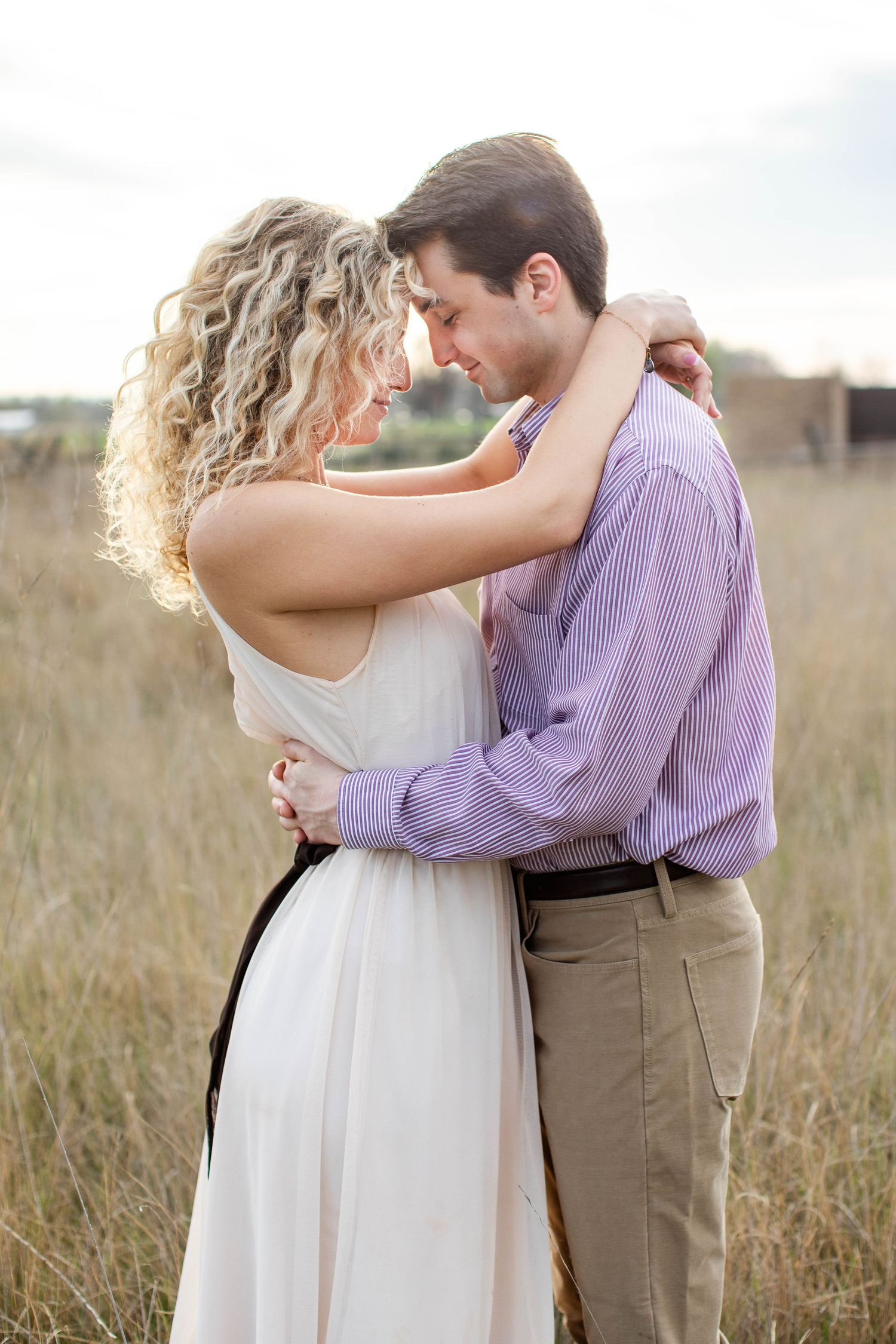 A couple embraces romantically in a golden field, woman in white dress with black sash, man in purple striped shirt and khakis.