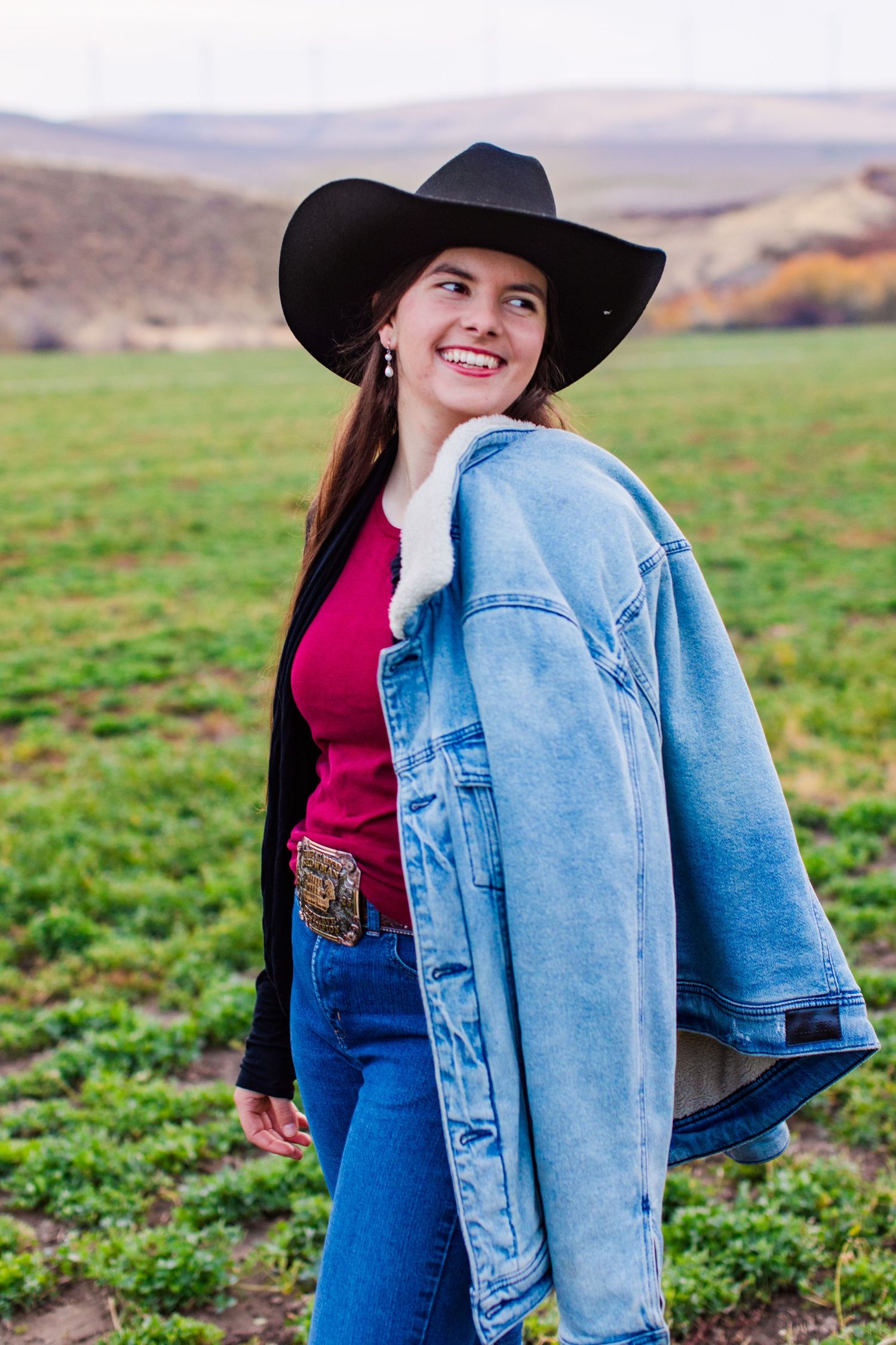 Woman in denim jacket and cowboy hat poses in front of green field and mountain landscape.