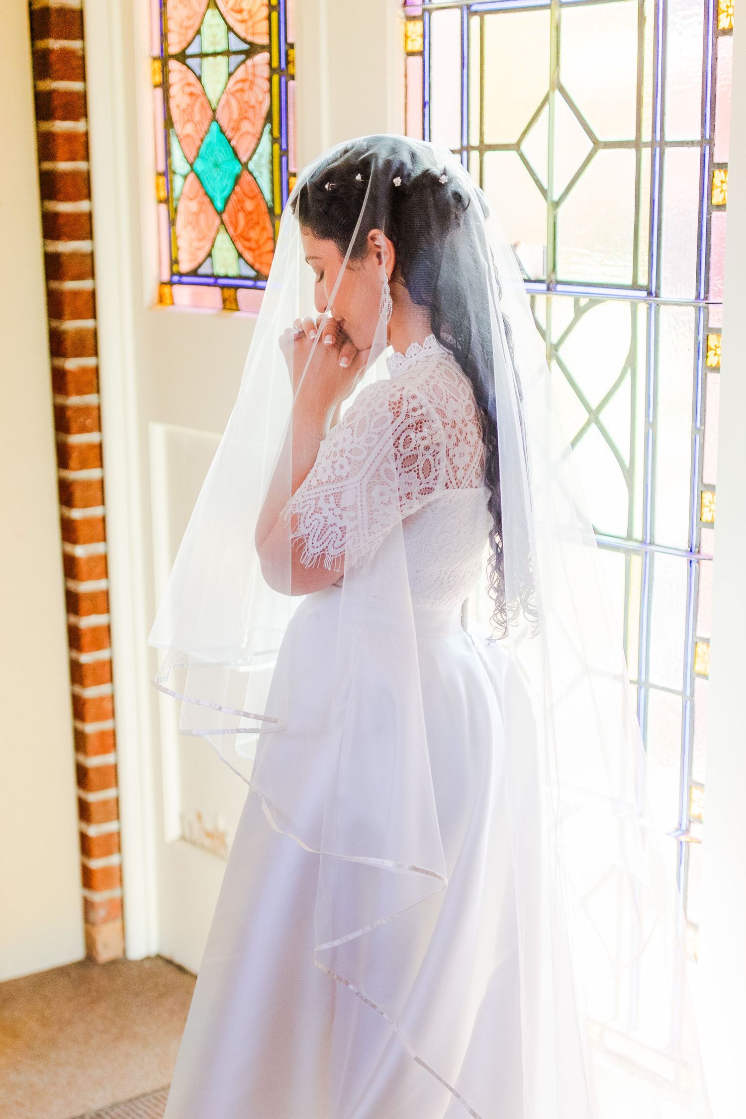 Bride in white lace gown photographed against stained glass church windows.