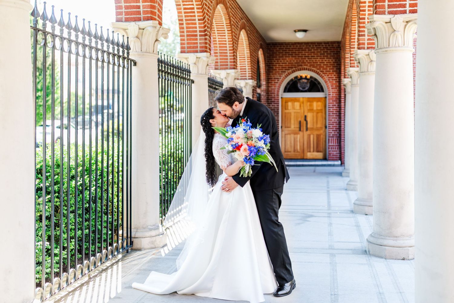 Wedding couple embracing in an elegant colonnade with iron fencing and brick architecture.