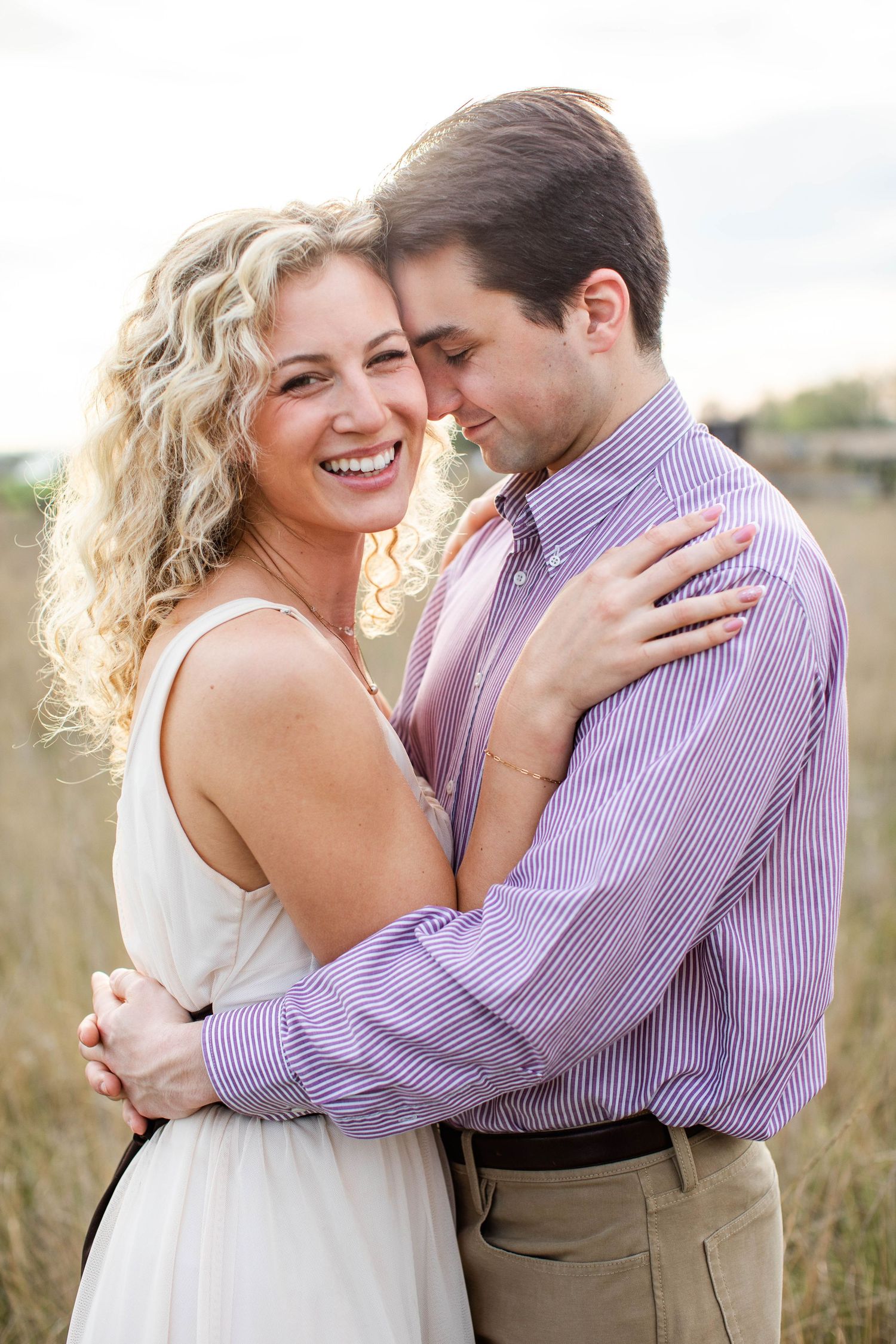 A smiling blonde woman in a white dress embraces a man in a purple striped shirt outdoors in a field.