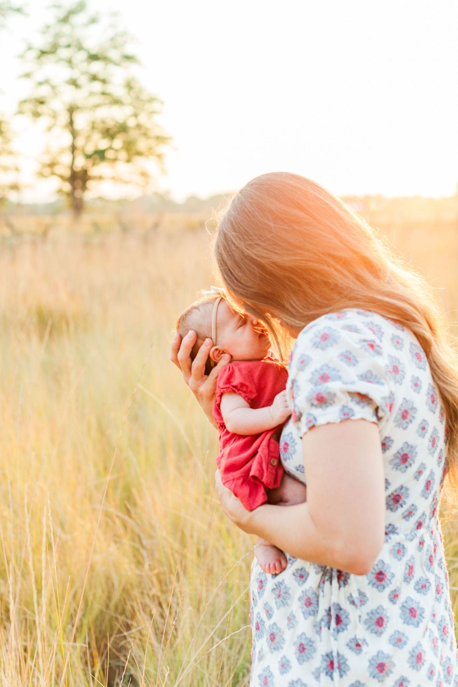 Mother holding child against golden sunset in grassy field wearing floral dress.
