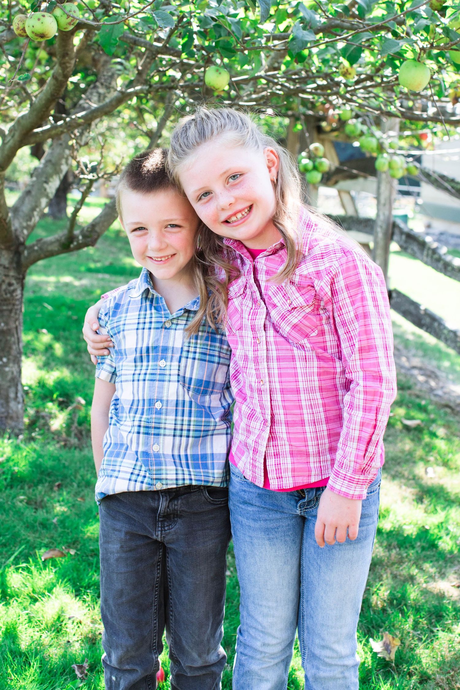 Children in plaid shirts pose together under green tree branches.