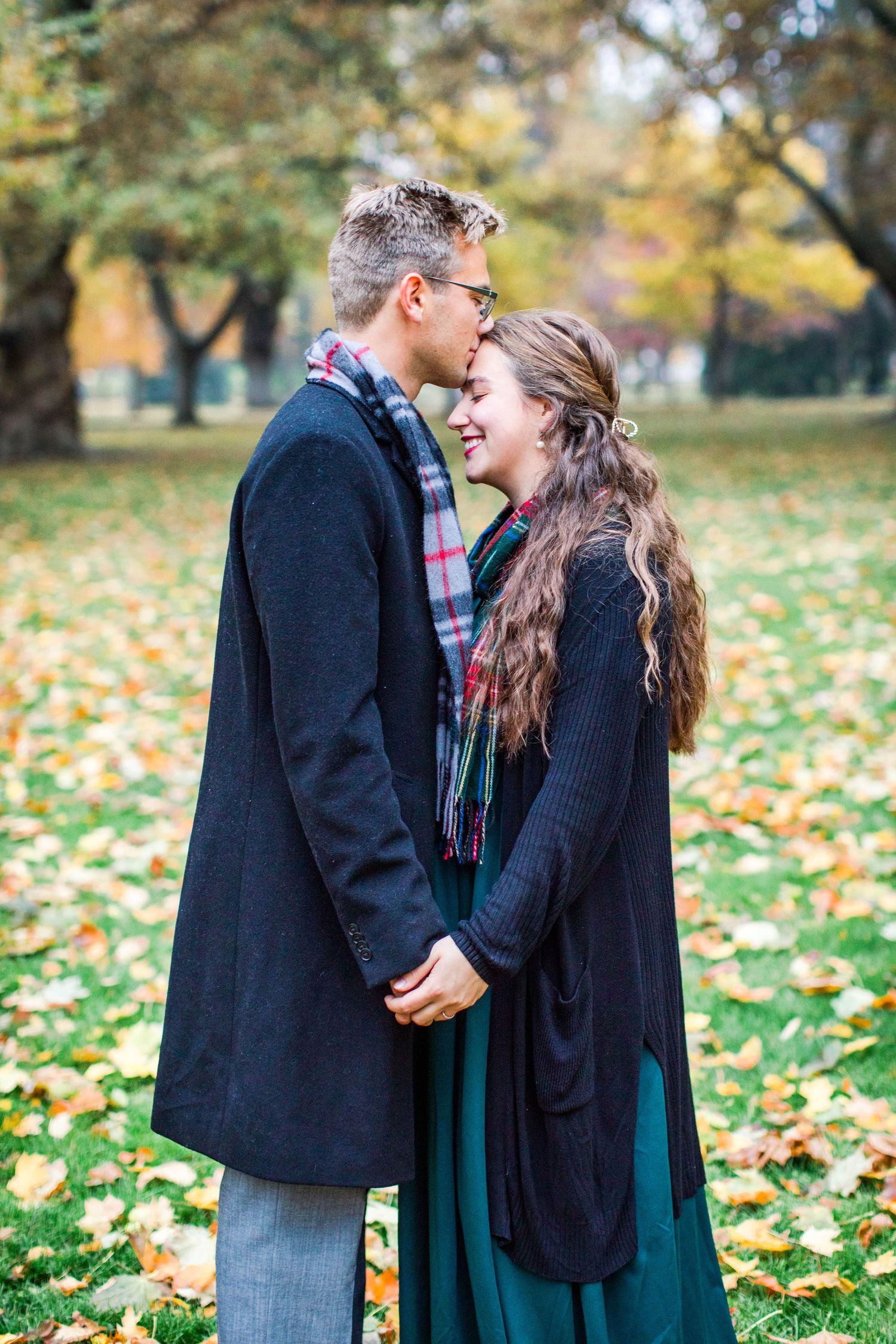 A couple shares romantic moments in a park filled with autumn leaves wearing dark coats.