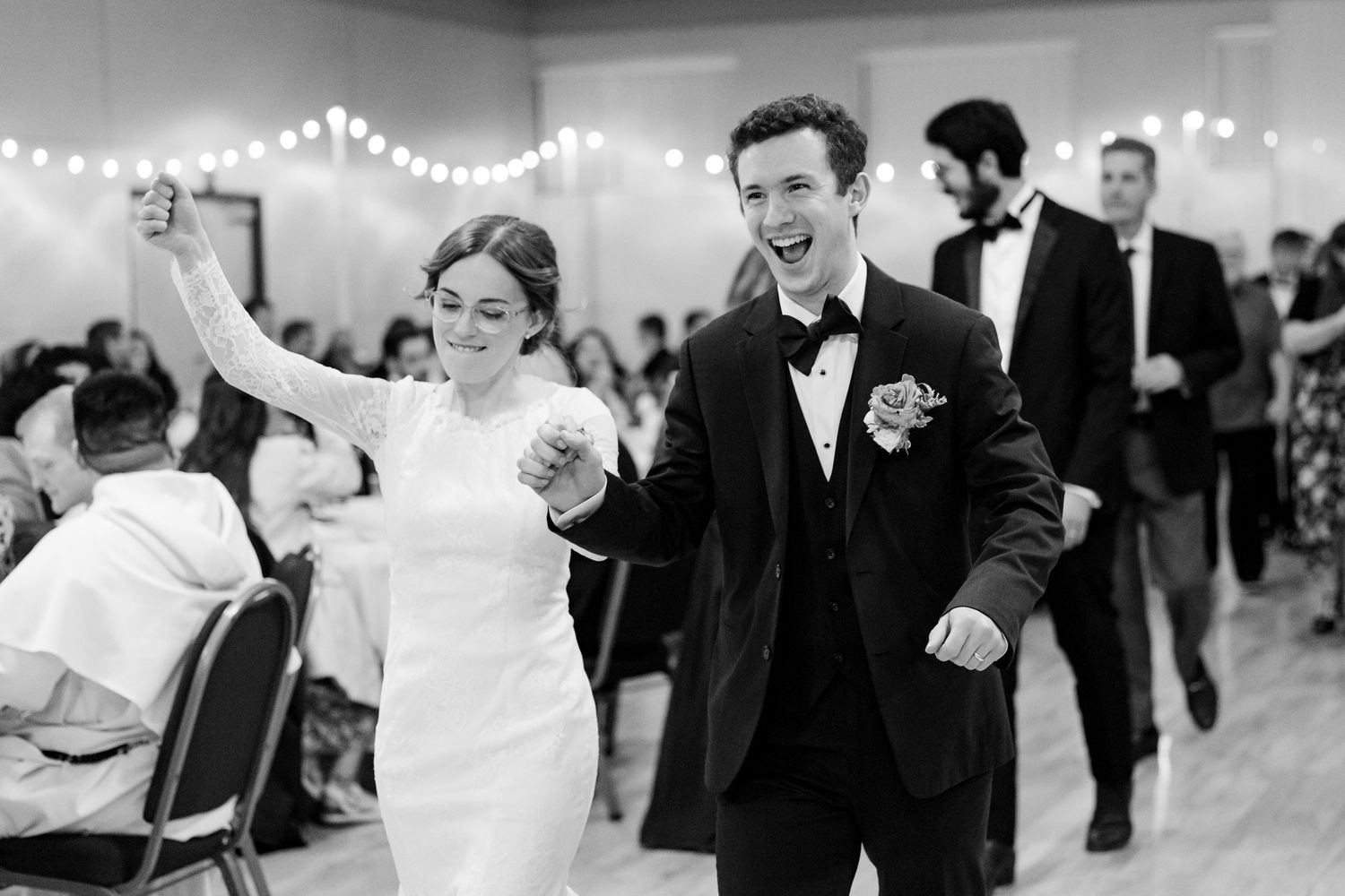 Black and white photo of a bride and groom joyfully entering their wedding reception, holding hands and smiling.