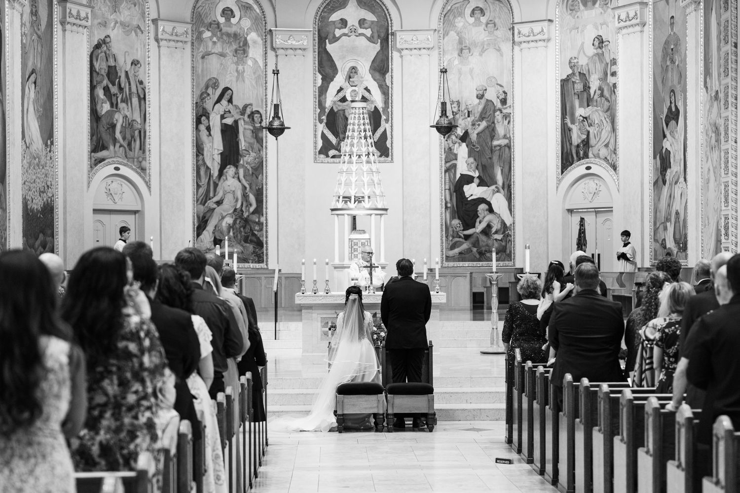 A black and white photograph of a wedding ceremony taking place inside an ornate church with religious murals on the walls.
