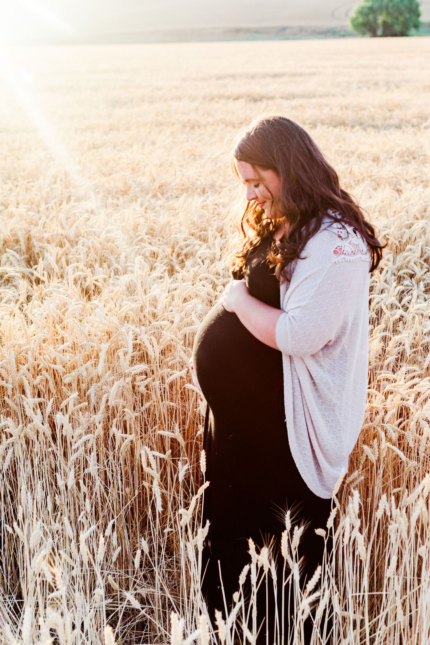 Side view of pregnant woman in white cardigan standing in golden wheat field at sunset.