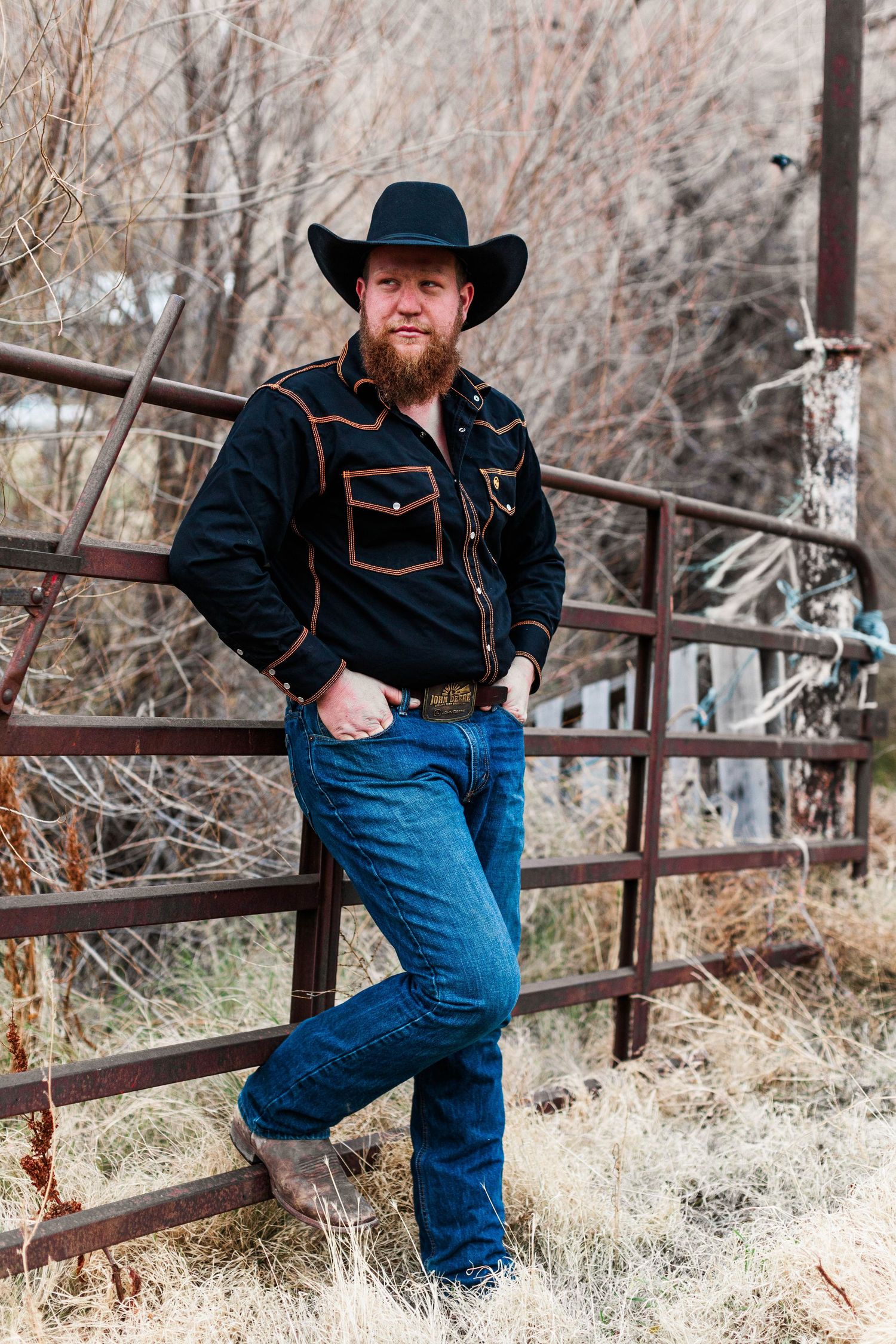 Person in western wear leans against rustic fence during winter photoshoot.