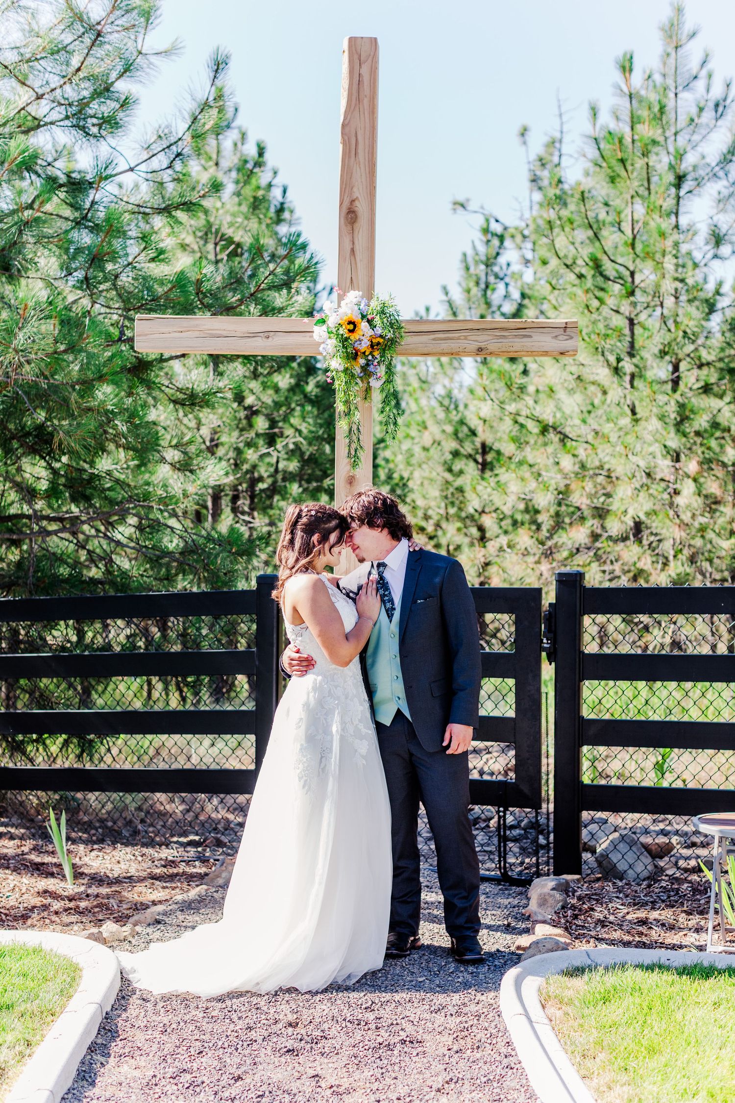Wedding couple embracing by wooden cross and black fence in outdoor garden setting.
