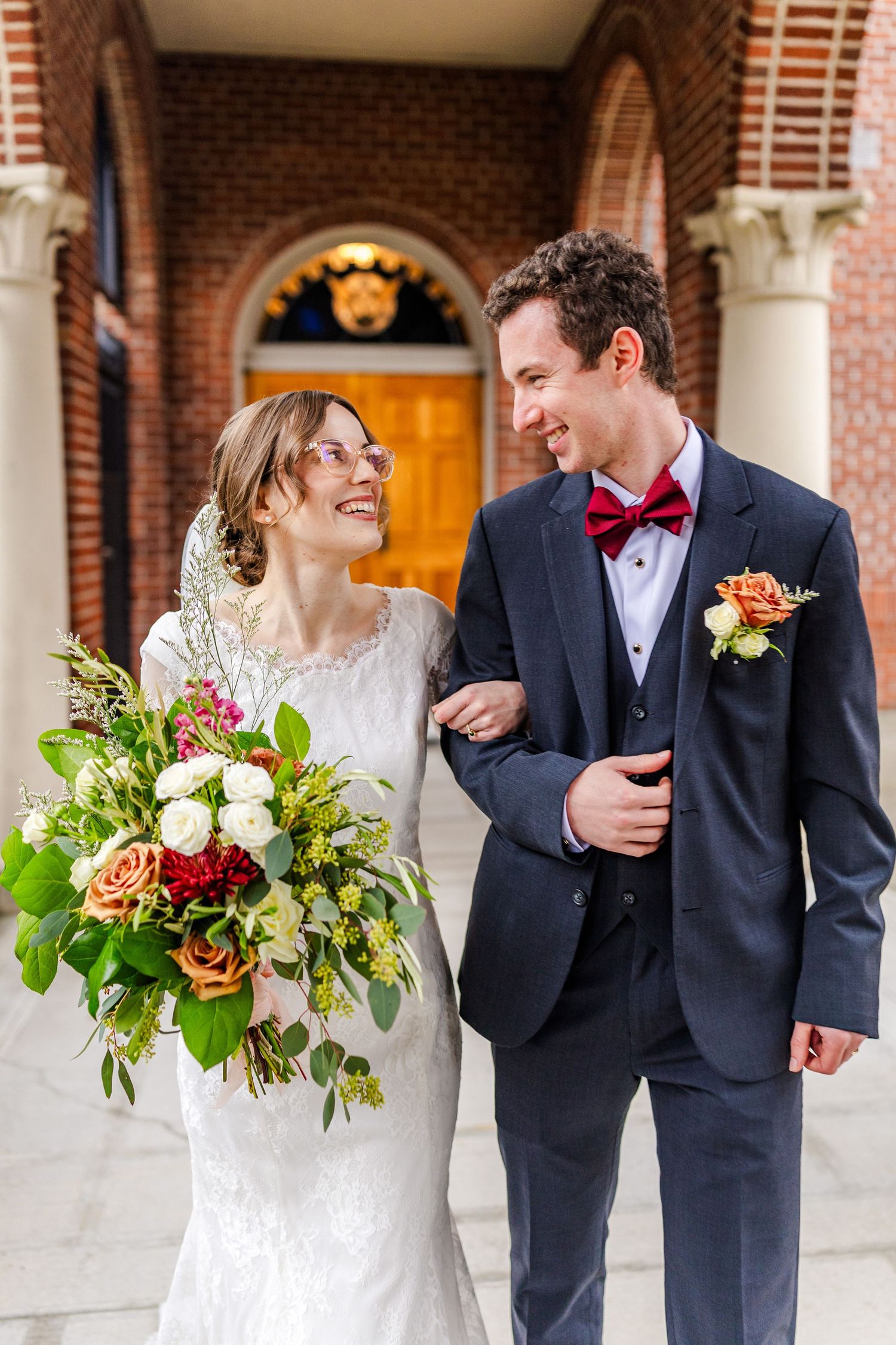 Newlyweds sharing intimate moments in formal attire with floral bouquets against brick architecture.
