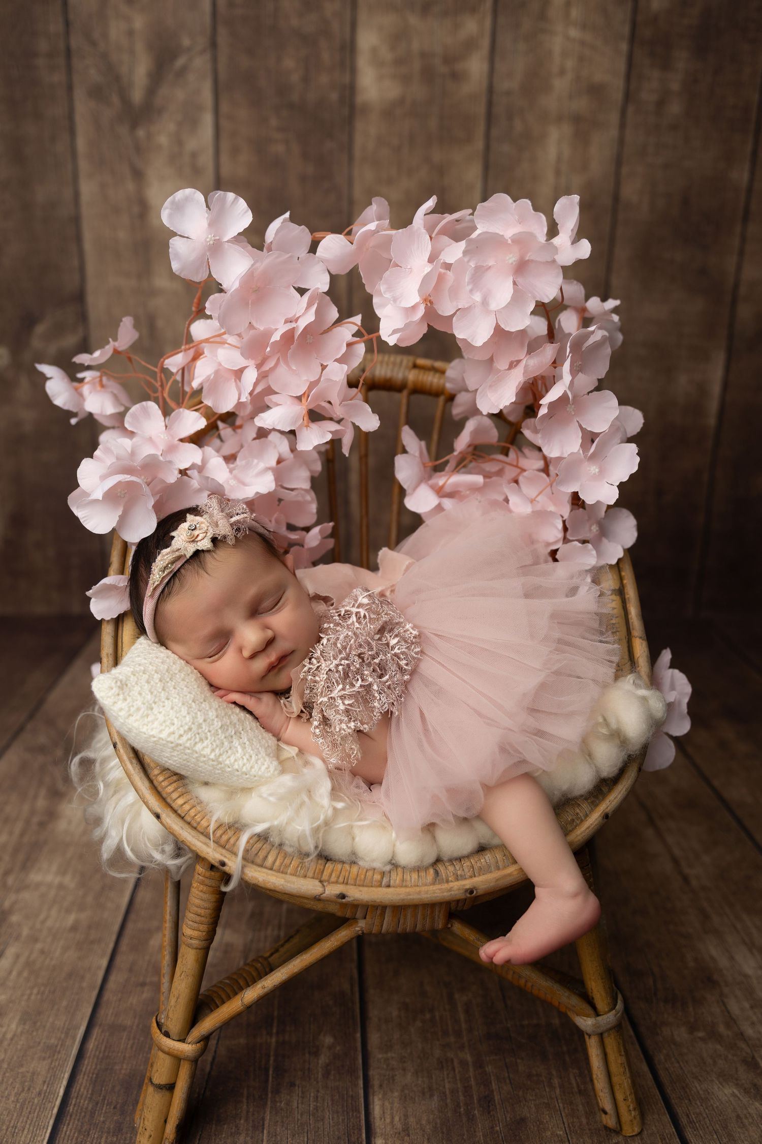 Newborn baby in pink tulle dress sleeping on rattan chair with floral decorations against rustic wooden backdrop.