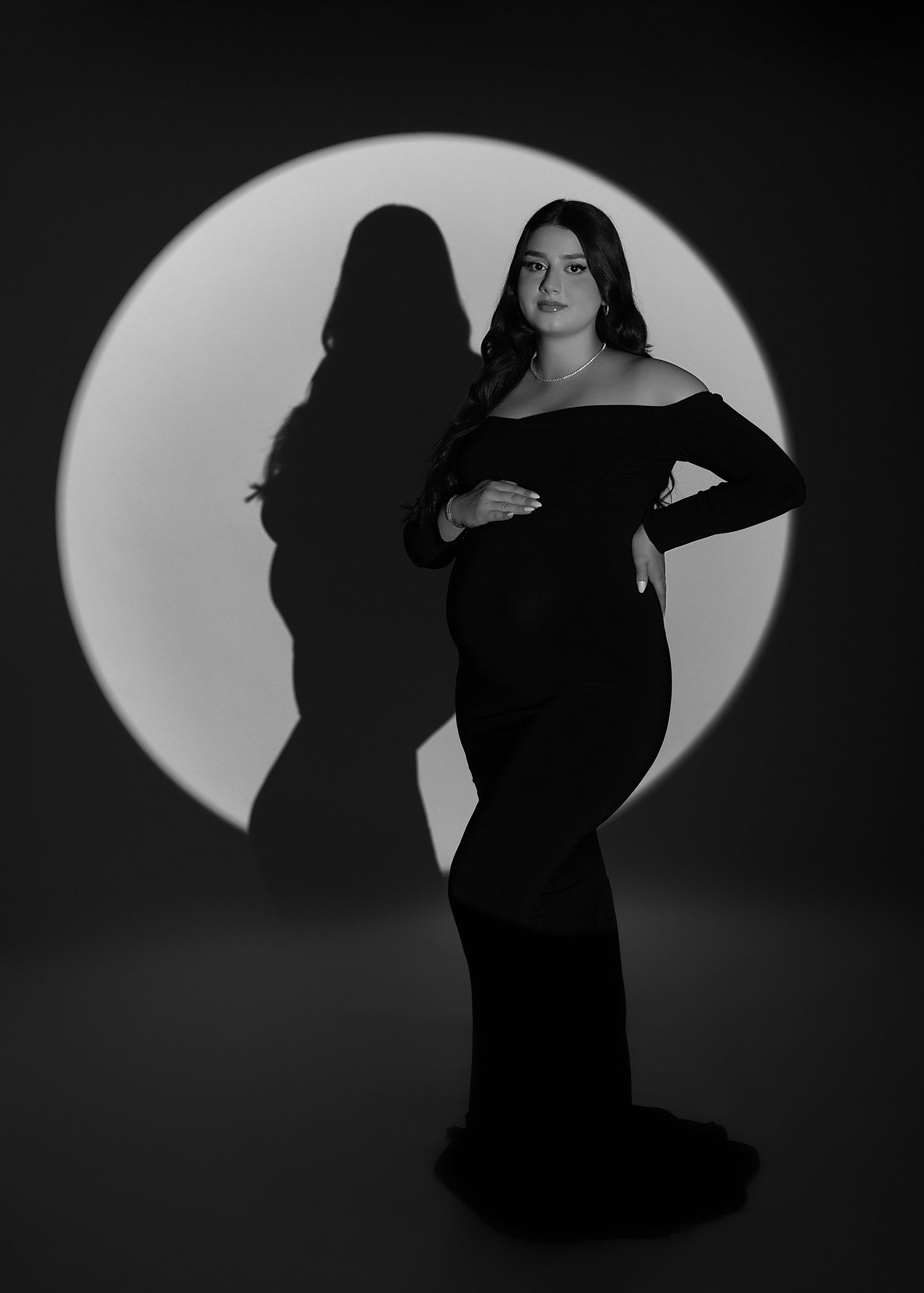 Black and white maternity photo of a woman in an elegant off-shoulder gown posed before a circular spotlight backdrop.