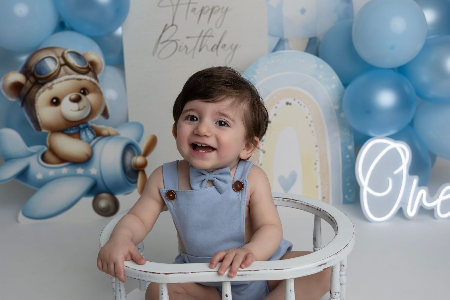 Baby boy in blue outfit smiling at first birthday photoshoot with teddy bear airplane backdrop and balloon arch.