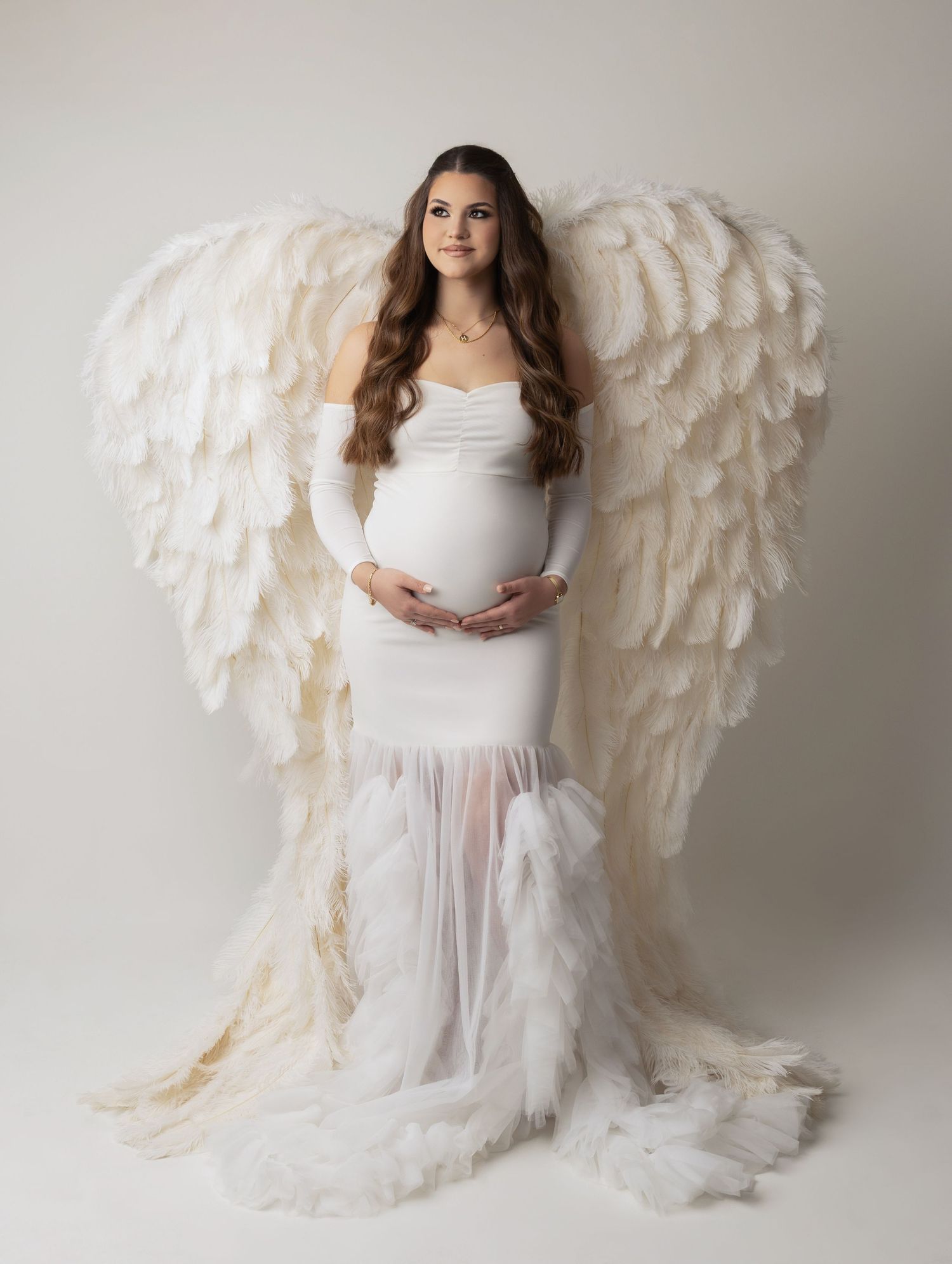Ethereal figure in flowing white dress and large feathered angel wings poses gracefully in studio photoshoot series.