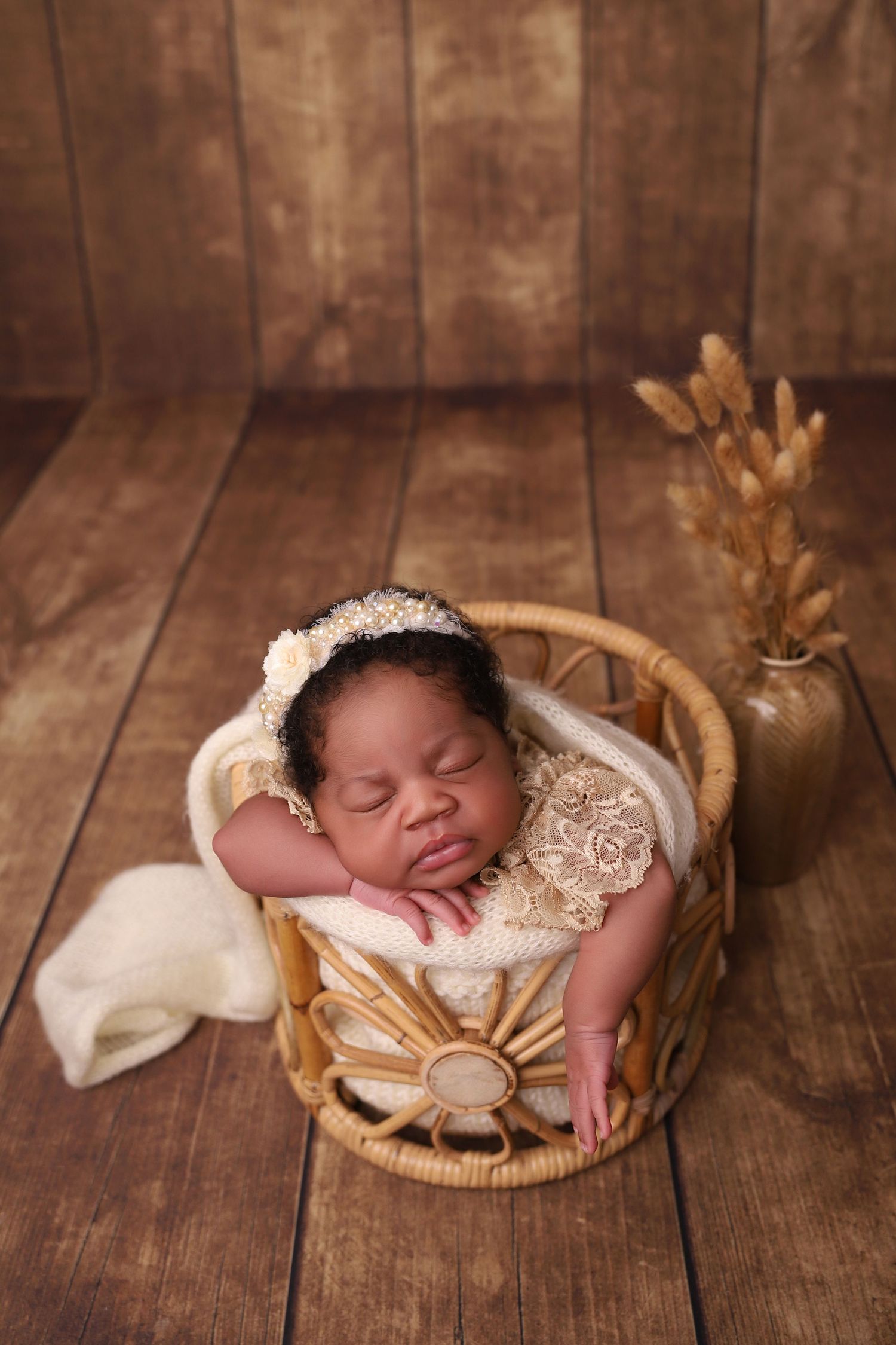 Sleeping newborn rests peacefully in woven basket with lace wrap on rustic wooden floor with dried flowers in background.
