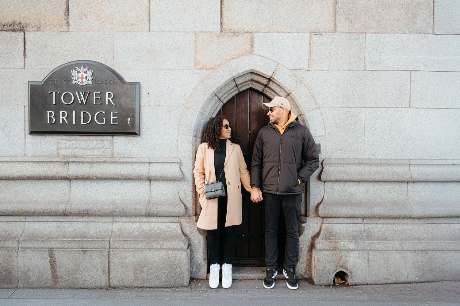 Couple stands in an arched stone doorway beneath a Tower Bridge sign in London.
