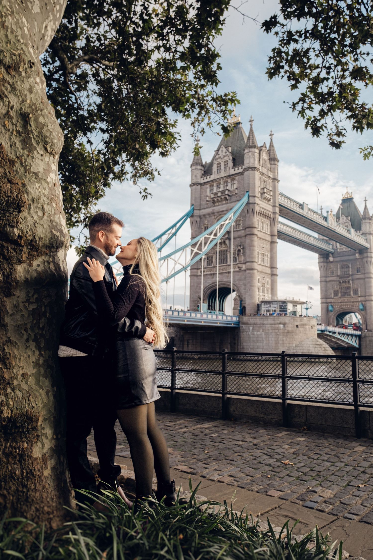 Romantic couple embracing near Tower Bridge in London on a cobblestone path.