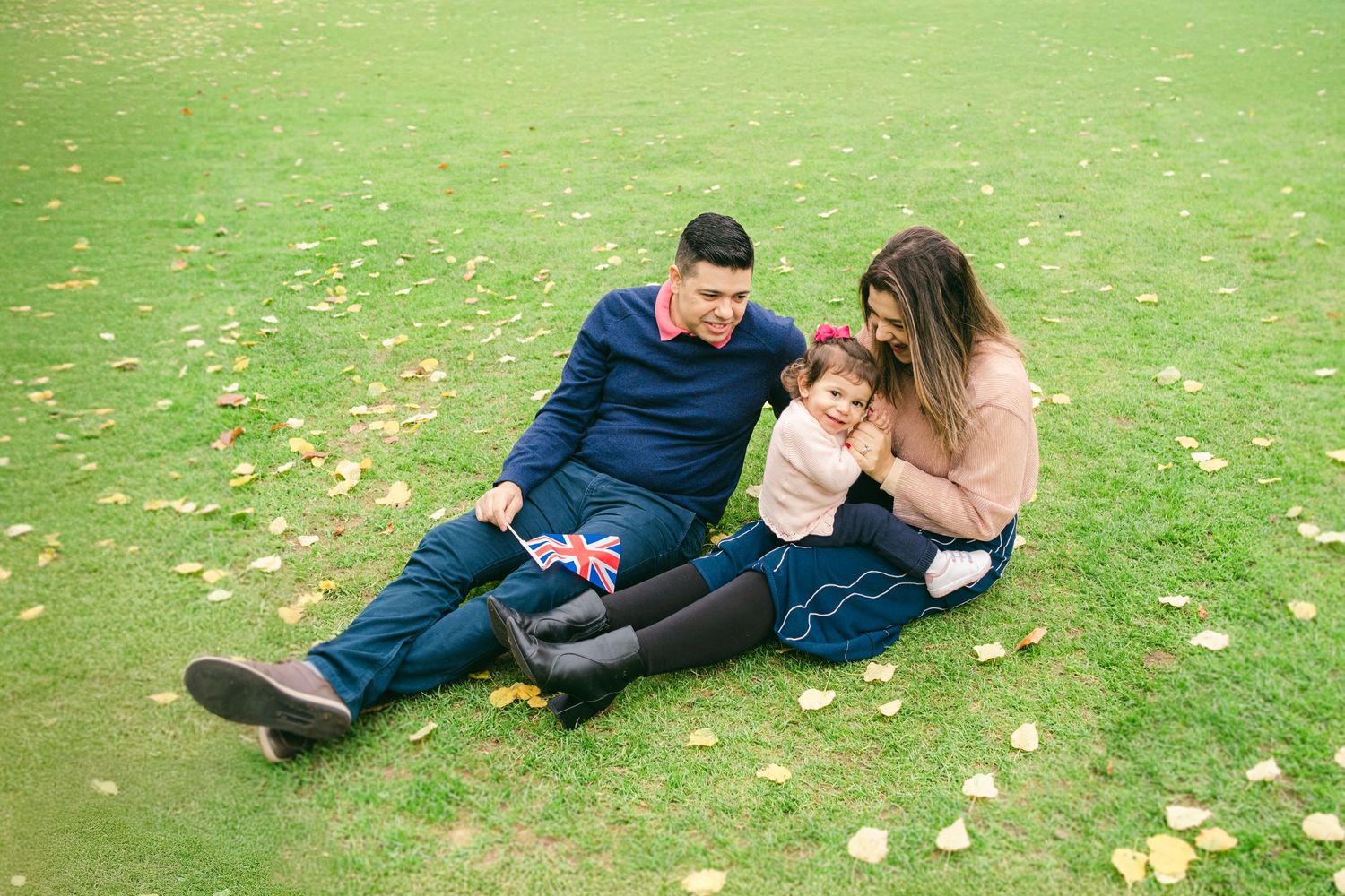 Family relaxing together on green grass covered with scattered autumn leaves.