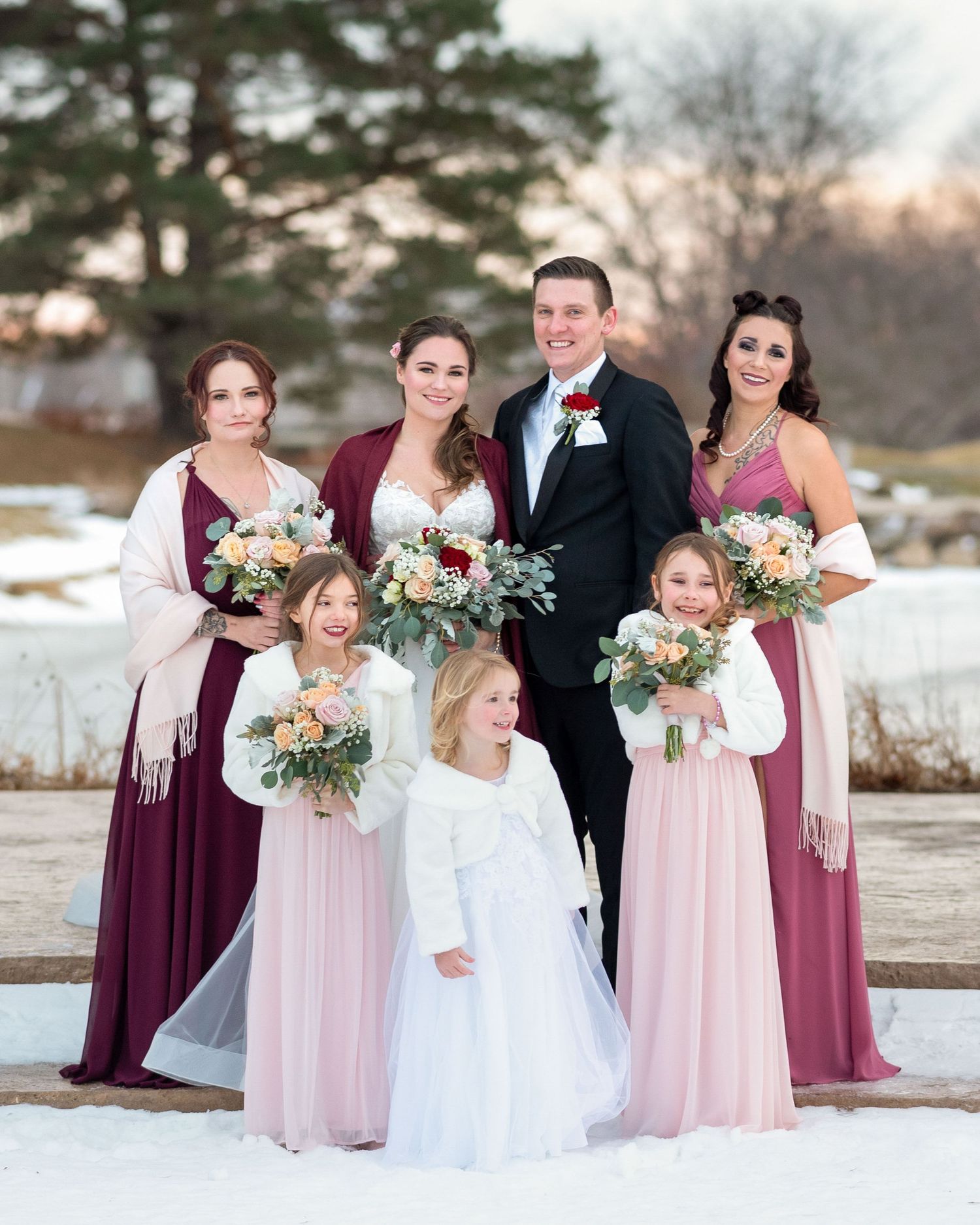 Wedding party poses in winter setting wearing burgundy and blush dresses with white floral bouquets.