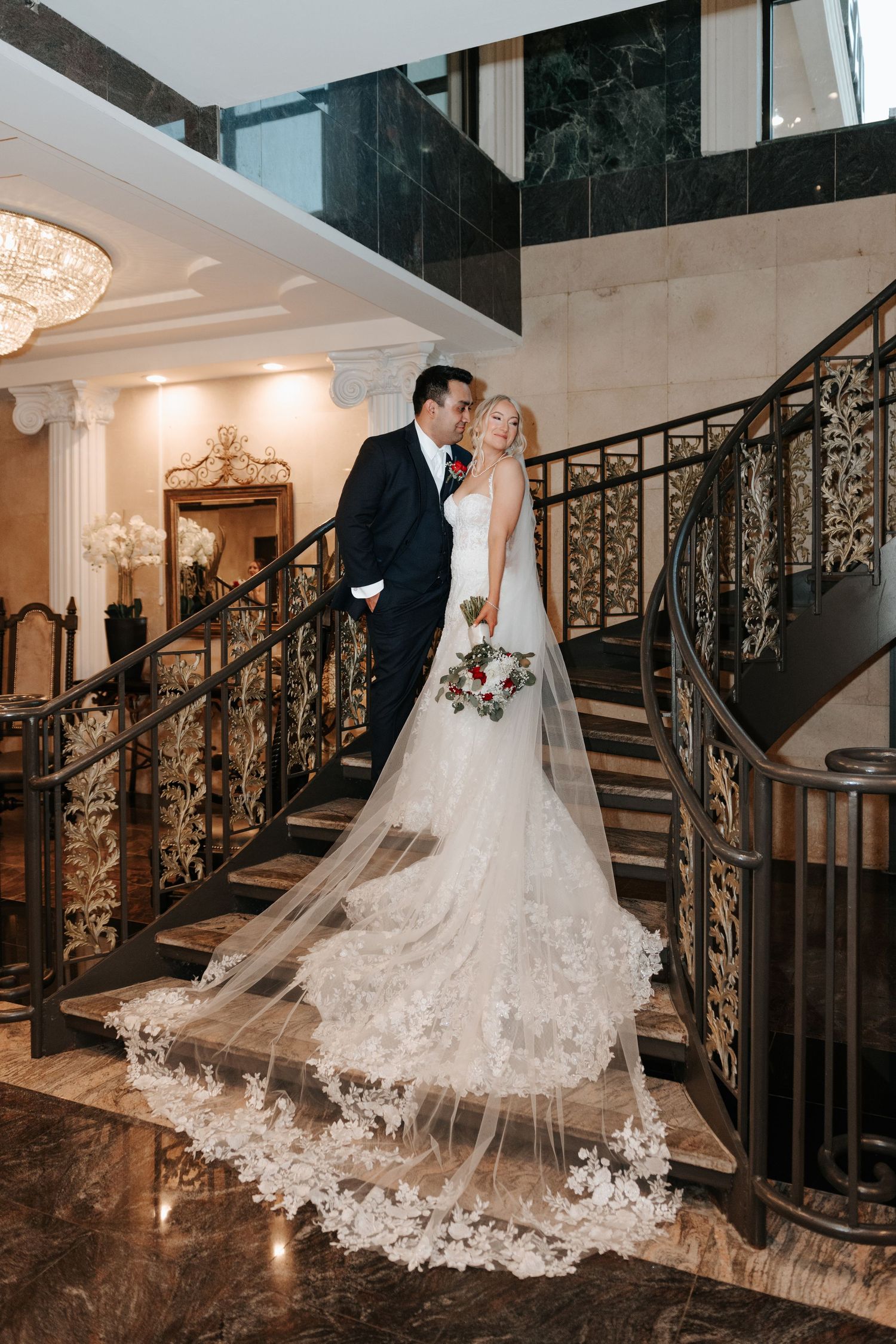 Wedding couple stands on ornate staircase with dramatic lighting and flowing cathedral veil.