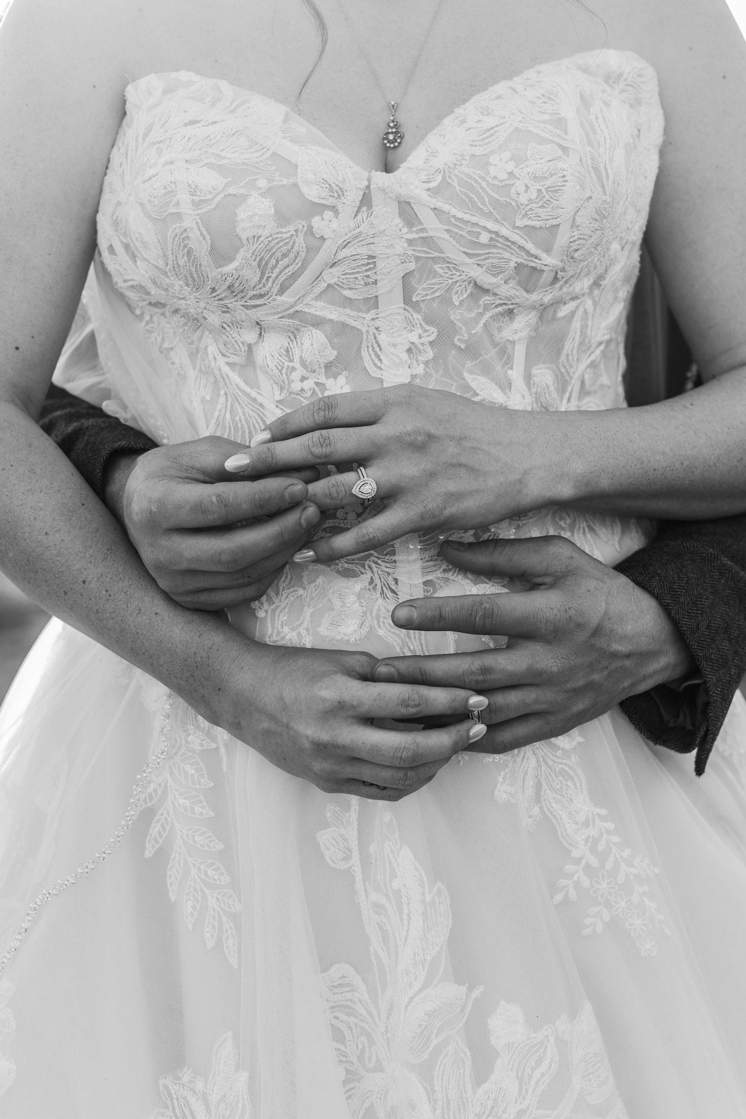 Black and white photo showing hands with rings embracing a white lace wedding dress bodice with delicate floral details.