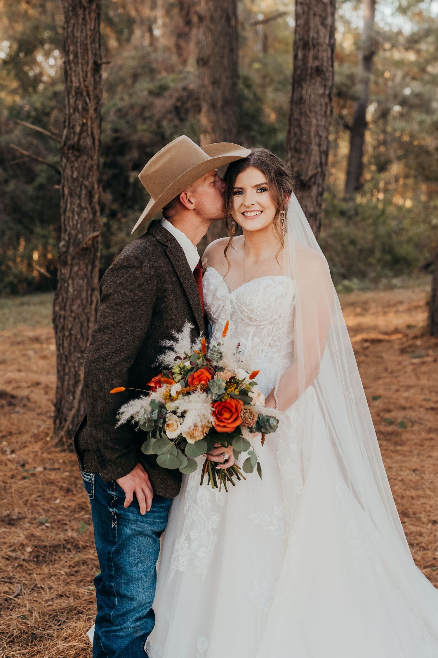 Romantic wedding couple embraces in a forest setting with autumn pine trees and a bridal bouquet of red and white roses.