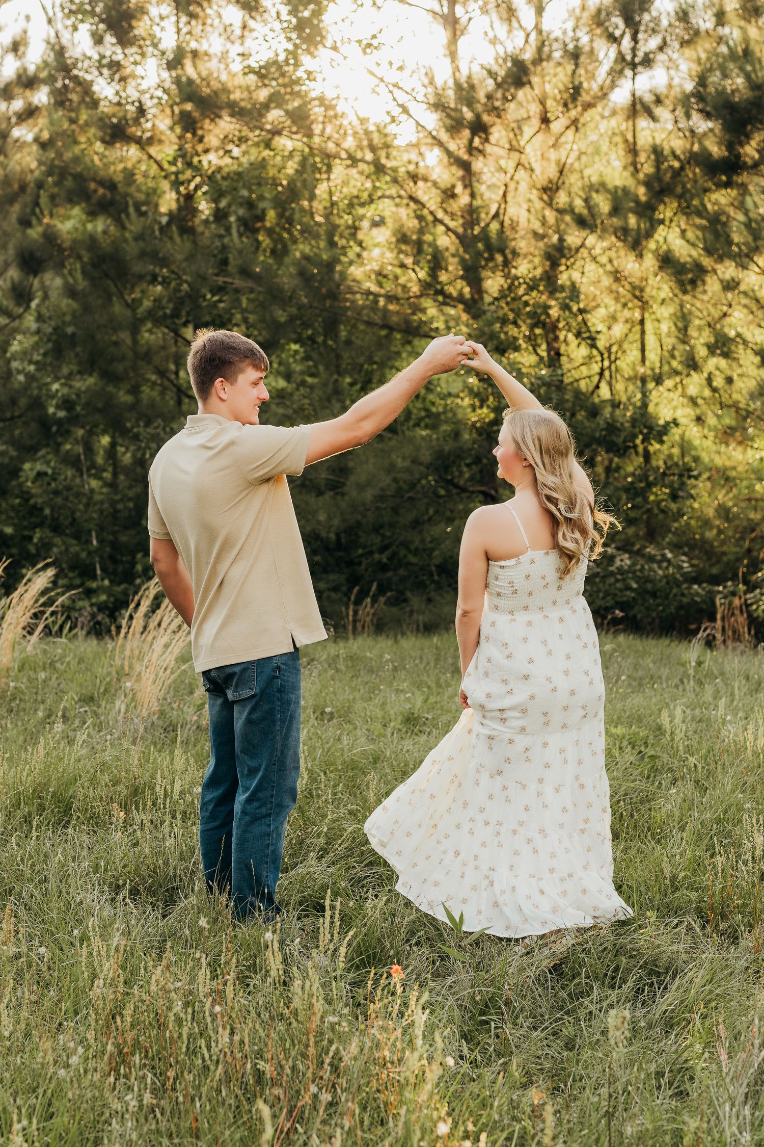 Couple dancing in a sunlit meadow, man spinning woman in floral dress, lush green trees in background.