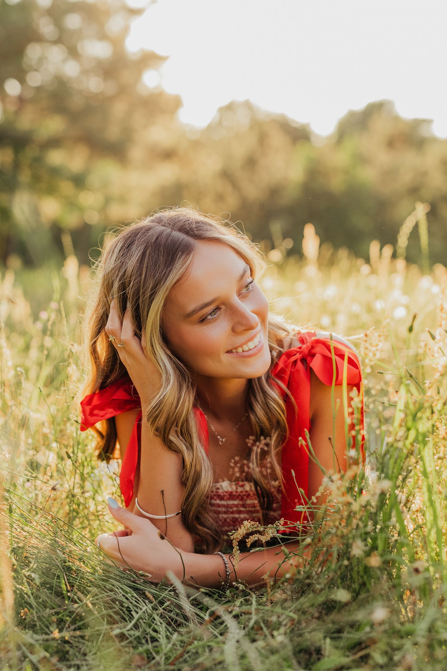 Young woman in red dress lying in sunny meadow grass, smiling warmly with golden backlight and wildflowers.