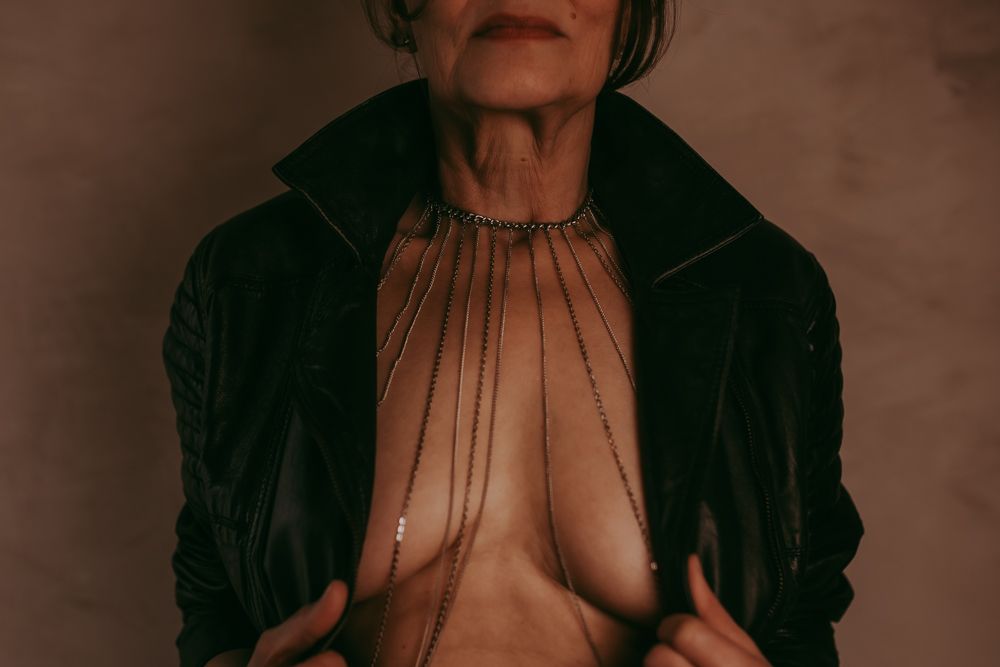 Elderly figure wearing a dark velvet jacket against brown backdrop with body art and jewelry visible.