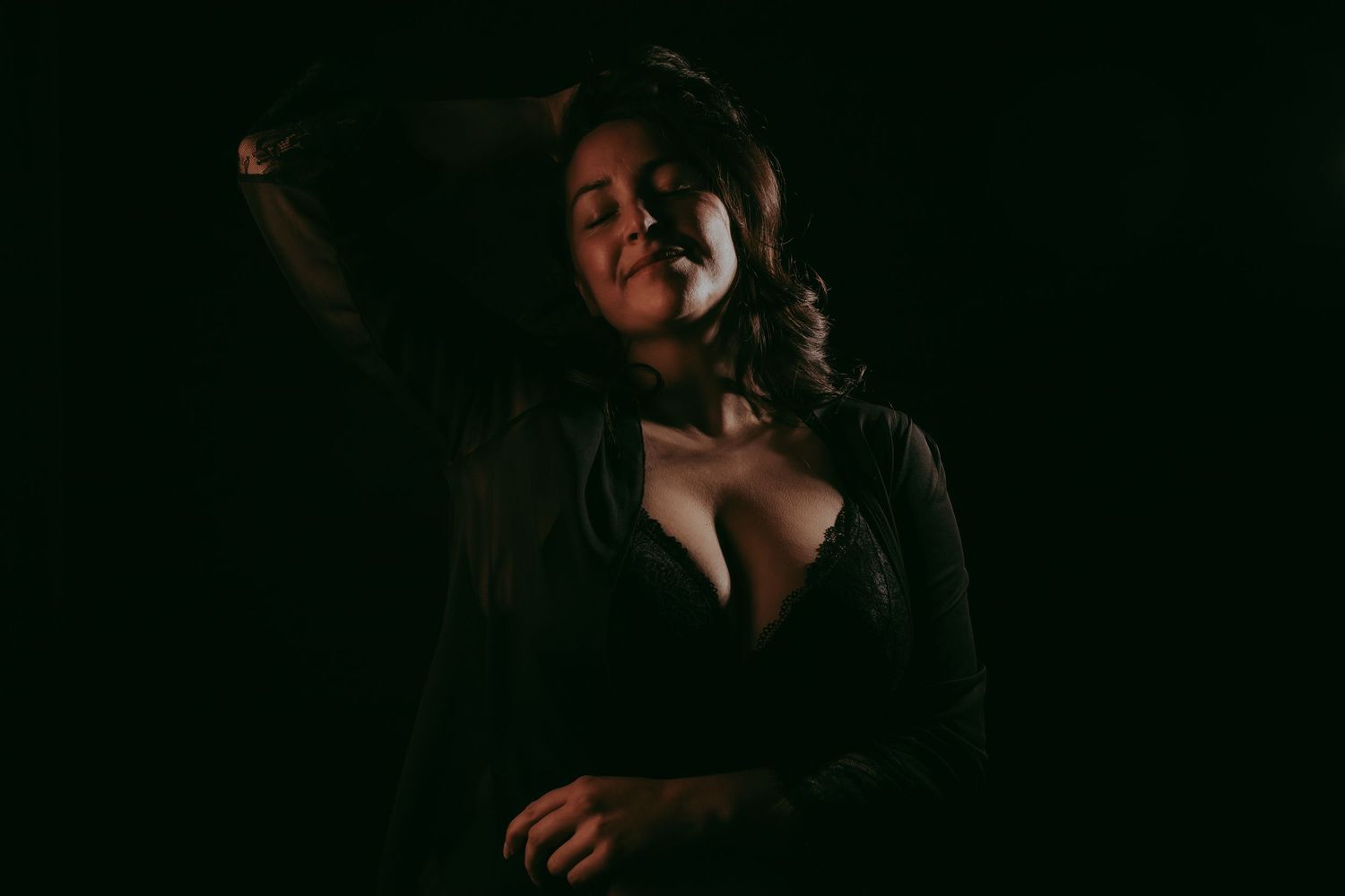 A woman poses dramatically in low-key lighting, wearing a dark jacket and lace top against a black background.
