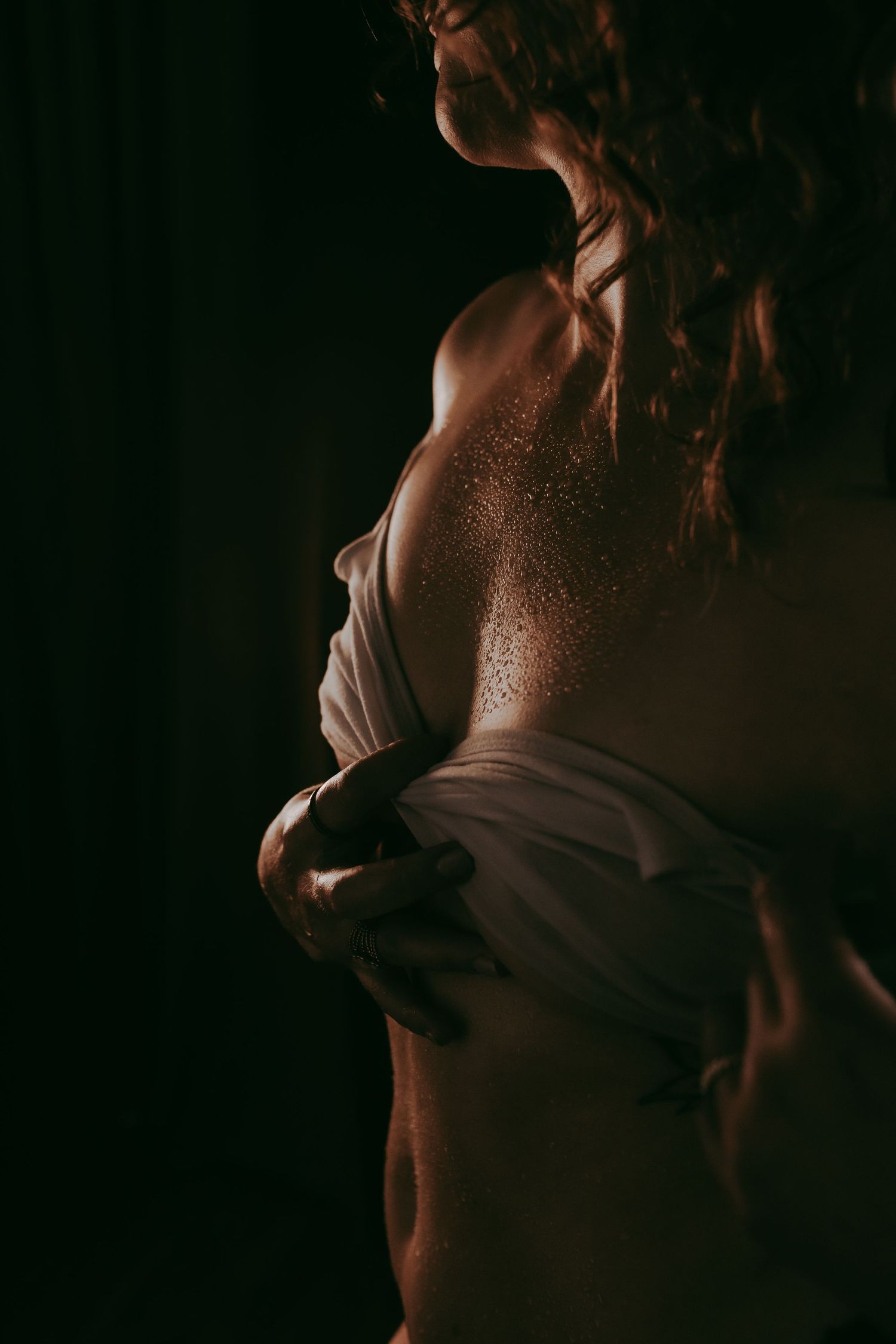A woman with curly hair holds white fabric against her body in dramatic low-key lighting with glittering skin.