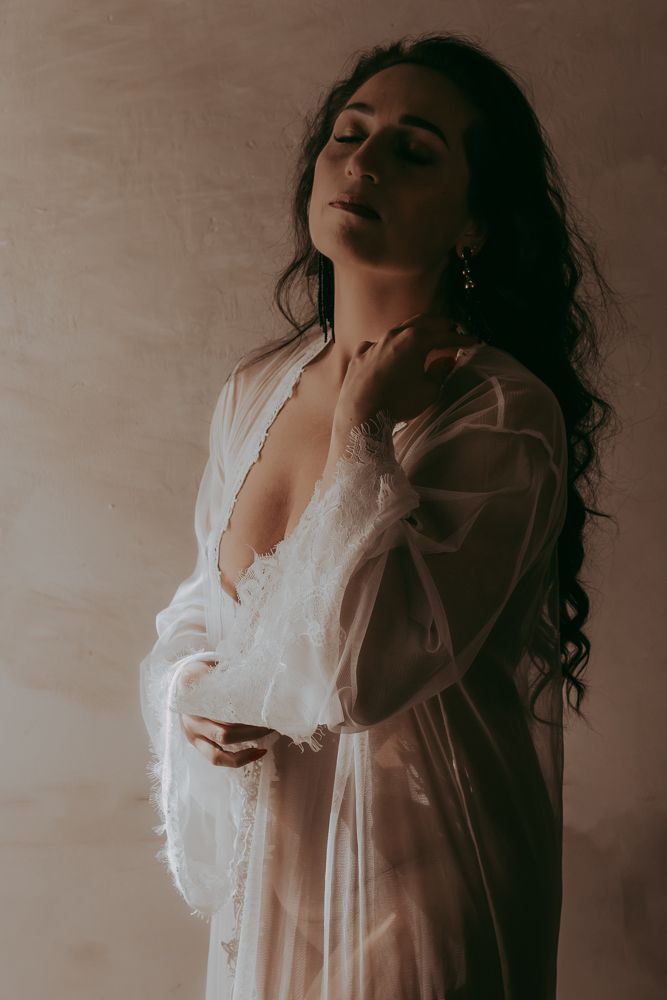 Ethereal portrait in sheer white dress with long dark curls and dramatic lighting against textured wall in moody atmosphere.
