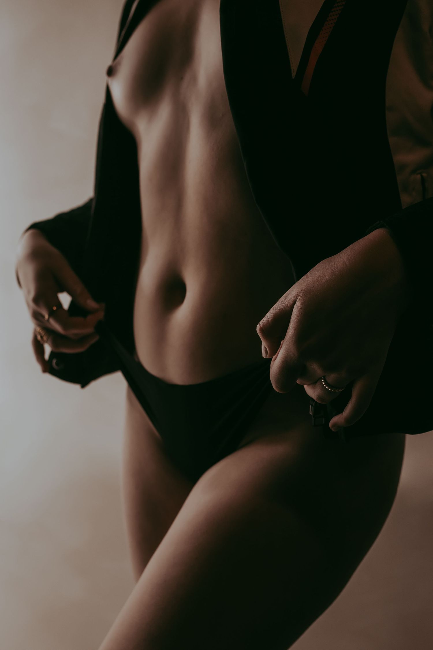 Woman in black lingerie and open blazer showcasing toned midriff, elegant rings on fingers.