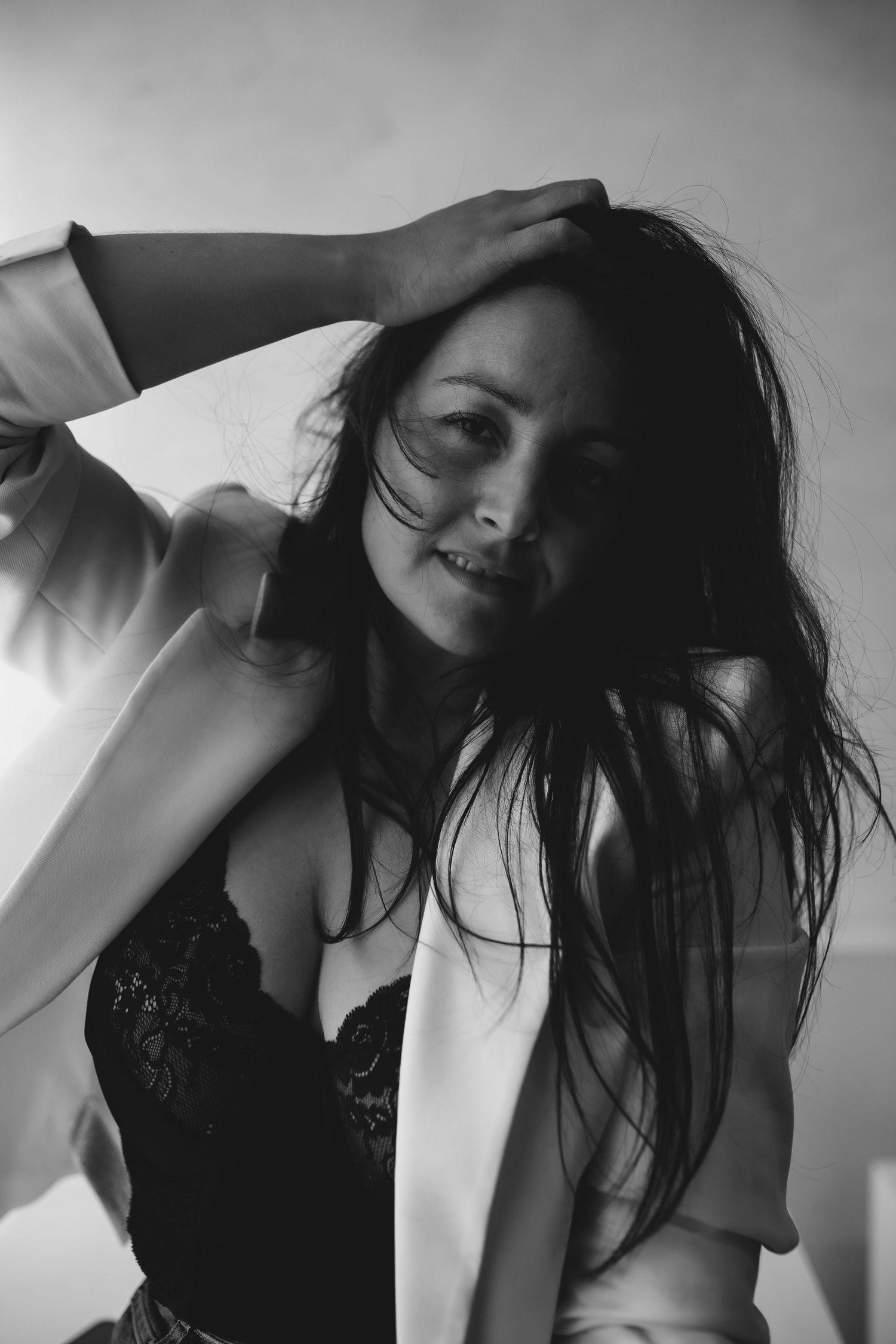 Black and white portrait of a woman with flowing dark hair, wearing lace top, hand in hair, smiling softly.