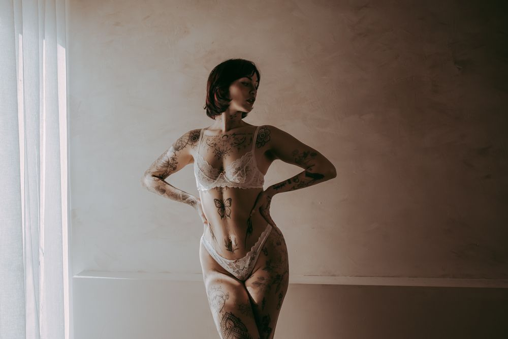 Artistic portrait of a figure in lingerie posing against neutral wall with dramatic side lighting and shadows.