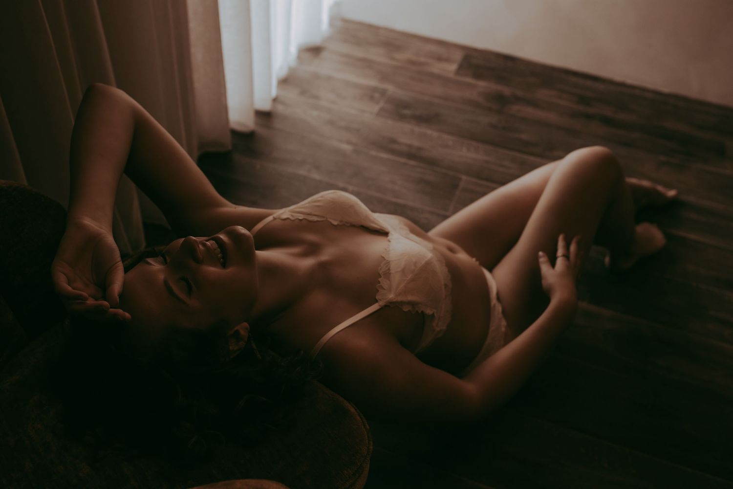 Woman in white lingerie posing on a dark wooden floor in moody, dimly lit boudoir photography.