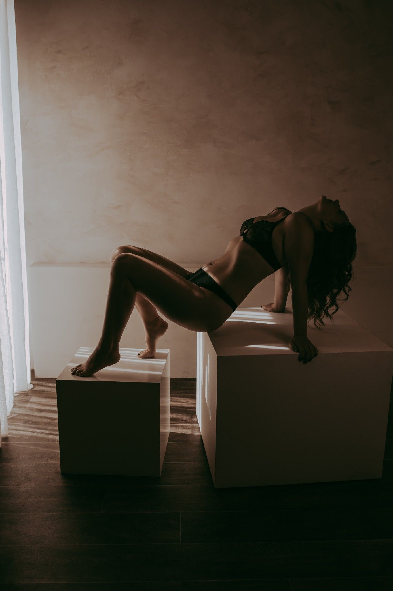 Boudoir foto van een vrouw die poseert op 2 witte blokken. Vrouw heeft een donker lingerie setje aan en het licht valt mooi naar binnen