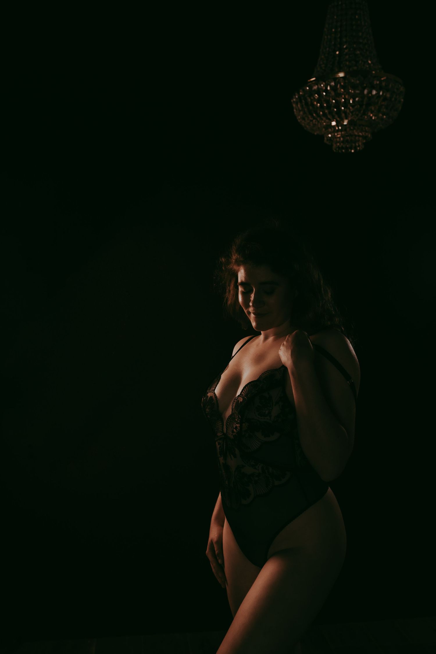 A woman in a dark lace bodysuit poses elegantly under a crystal chandelier in dramatic low-key lighting.
