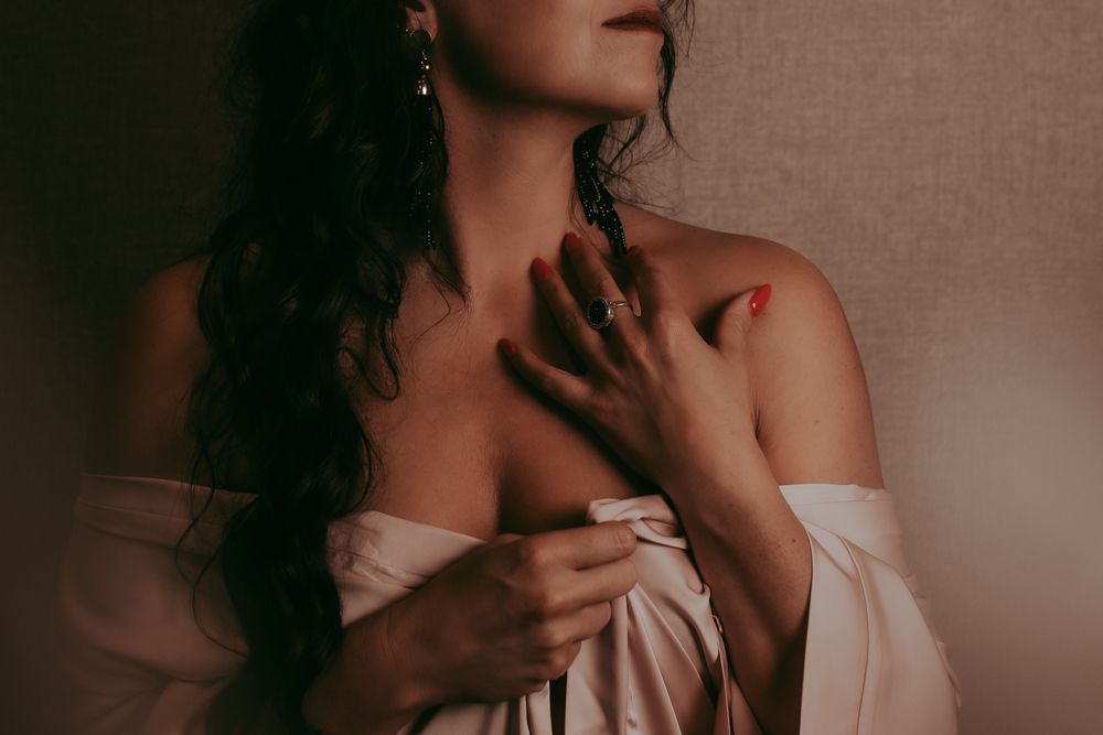 Moody boudoir photograph in warm tones showing figure in draped fabric with long dark wavy hair against neutral background.