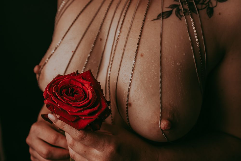 Artistic boudoir photograph showing a dark red rose being held against skin in moody lighting.
