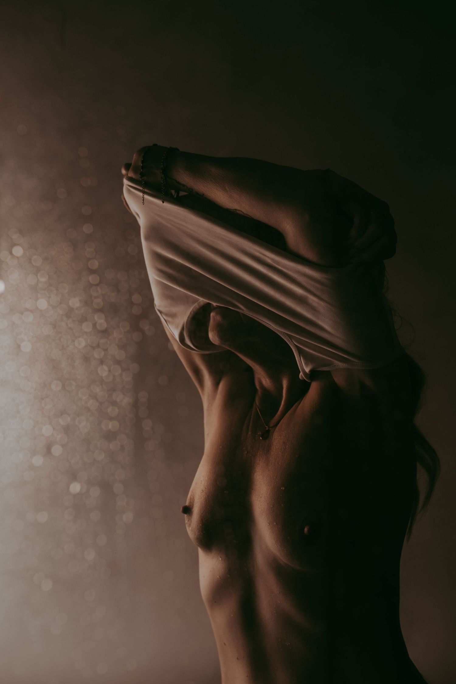 Artistic moody photograph of a person removing a white shirt, captured in dramatic low-key lighting with bokeh background.