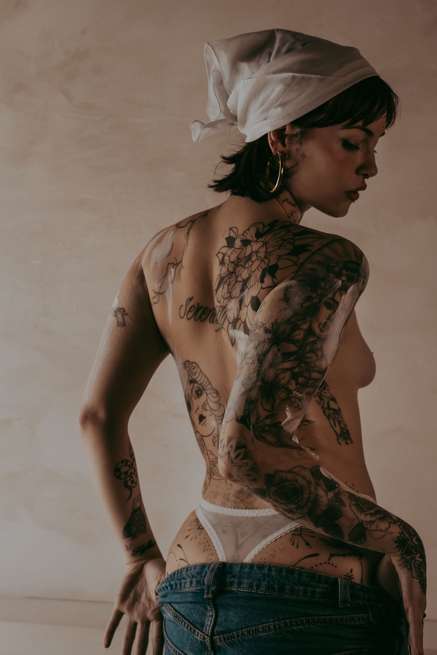 Boudoirfoto vrouw met tattoos en een wit doekje op haar hoofd zoals een boerin en met een zakkende spijkerbroek en wit verf op haar lichaam