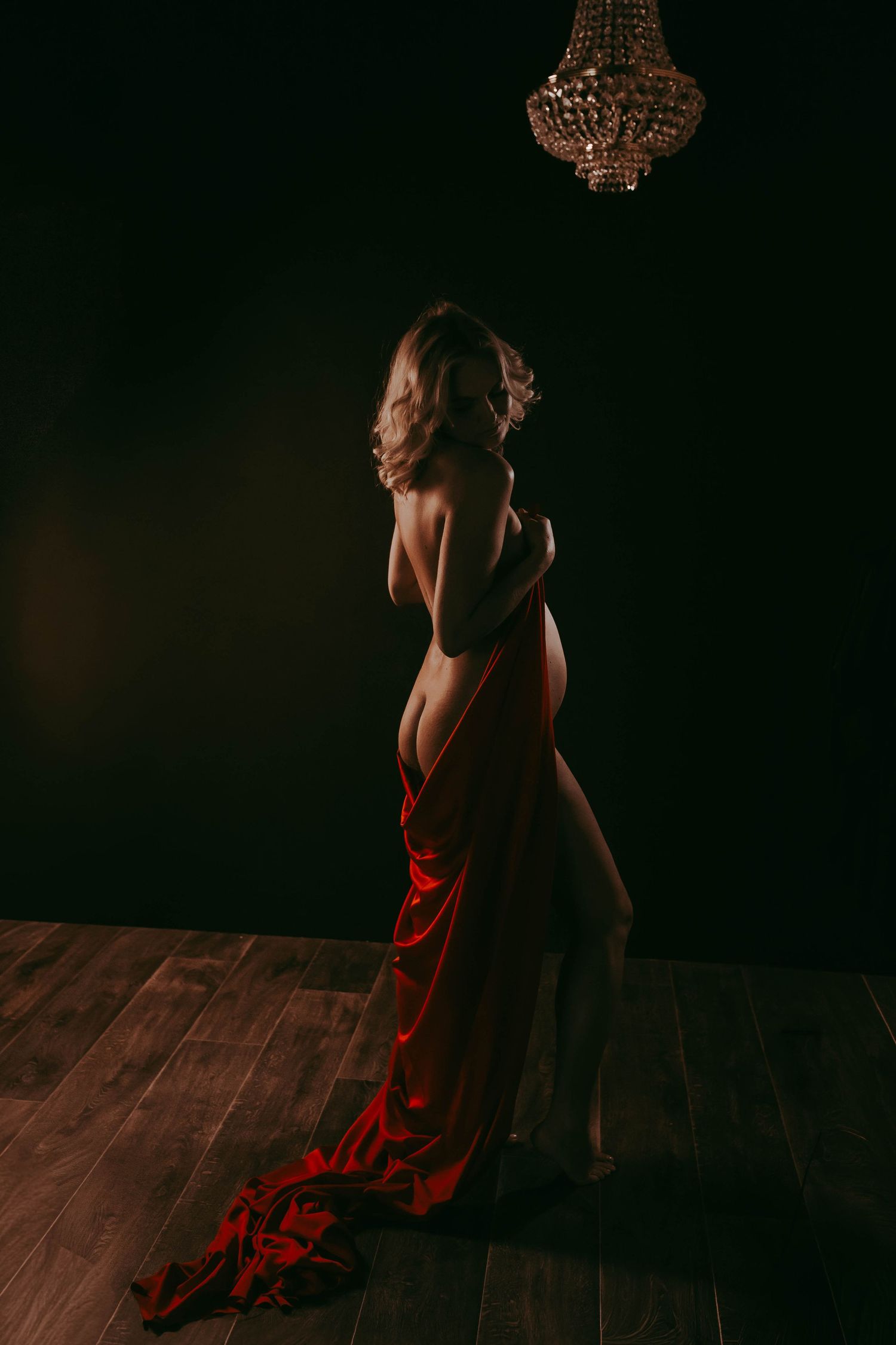 Elegant woman draped in red fabric standing in dramatic dark studio lighting beneath a crystal chandelier.
