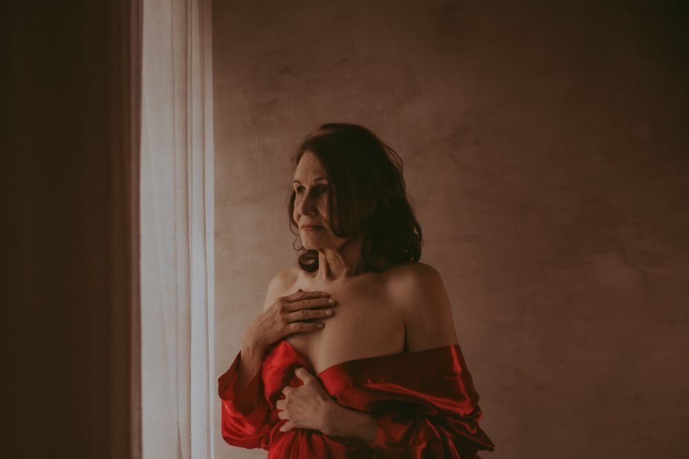 Moody portrait of person wearing red clothing by window with dramatic lighting and warm brown tones in background.