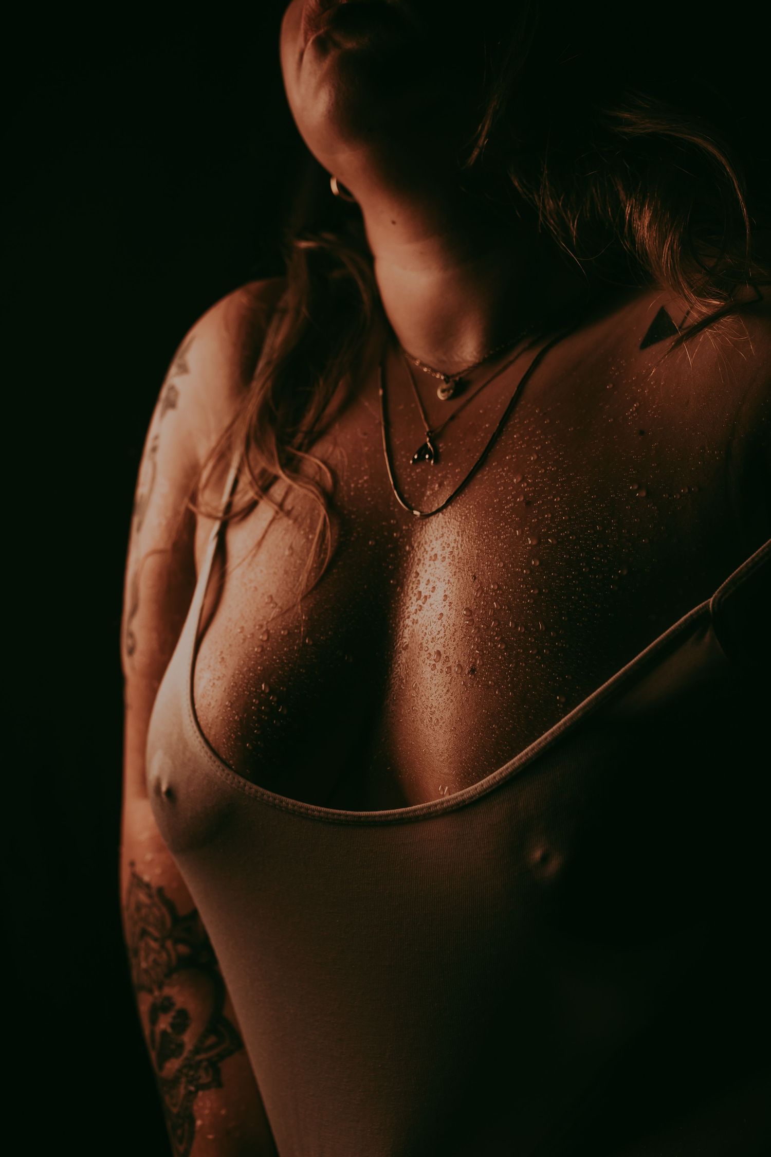 Woman with tattoos wearing layered necklaces and a low-cut top, glistening skin against dark background.
